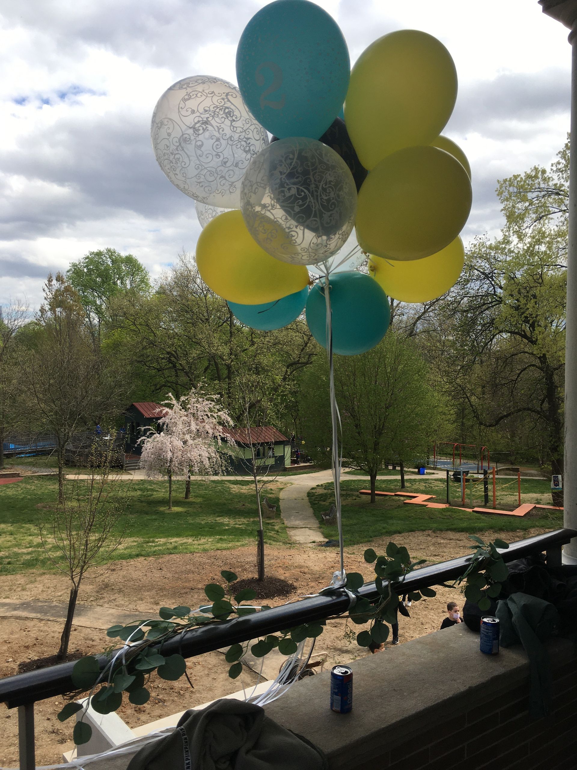 Cluster of teal, yellow, and silver balloons floating on a balcony overlooking a park