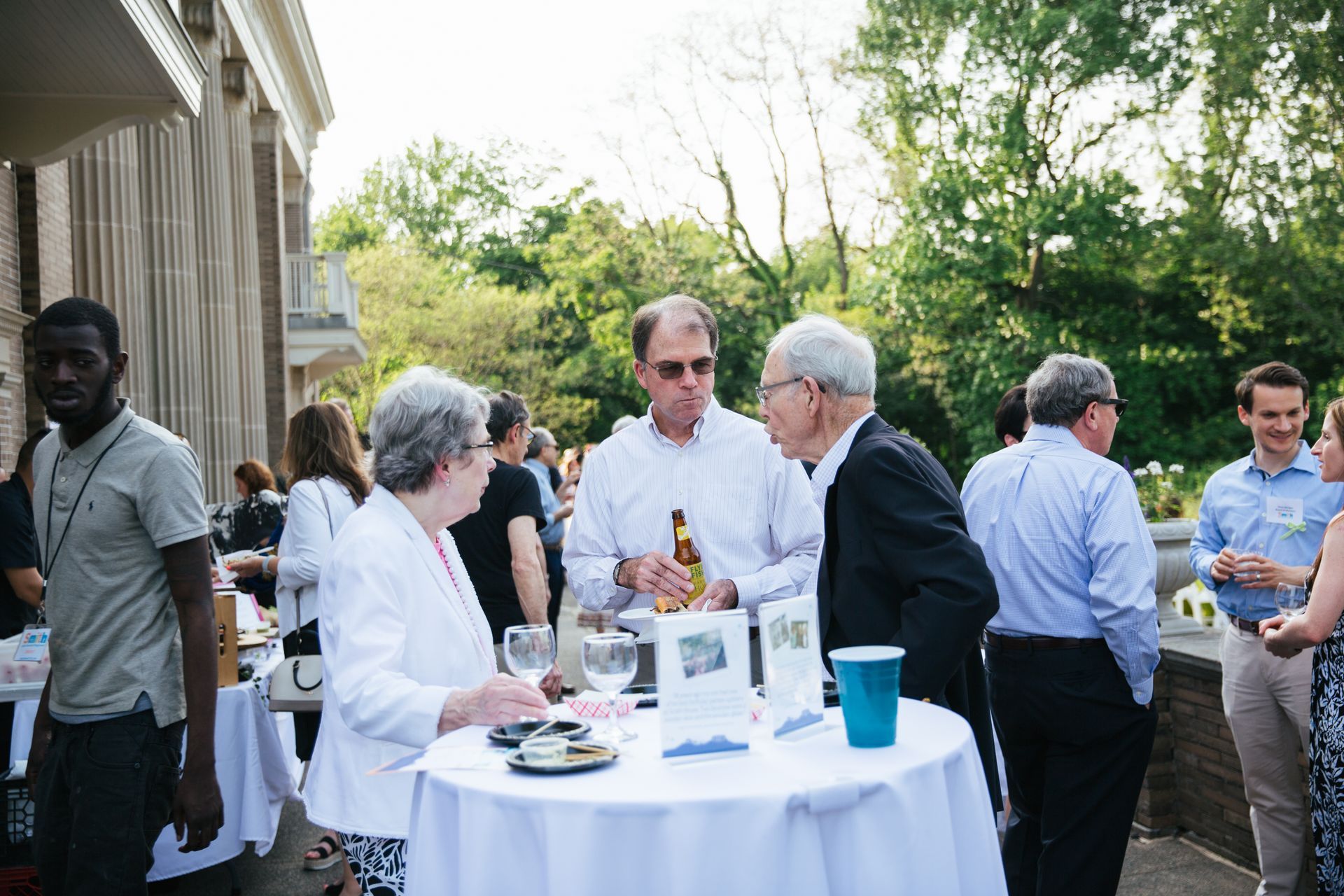 People mingling at an outdoor reception beside a building, with drinks and a round table in the foreground