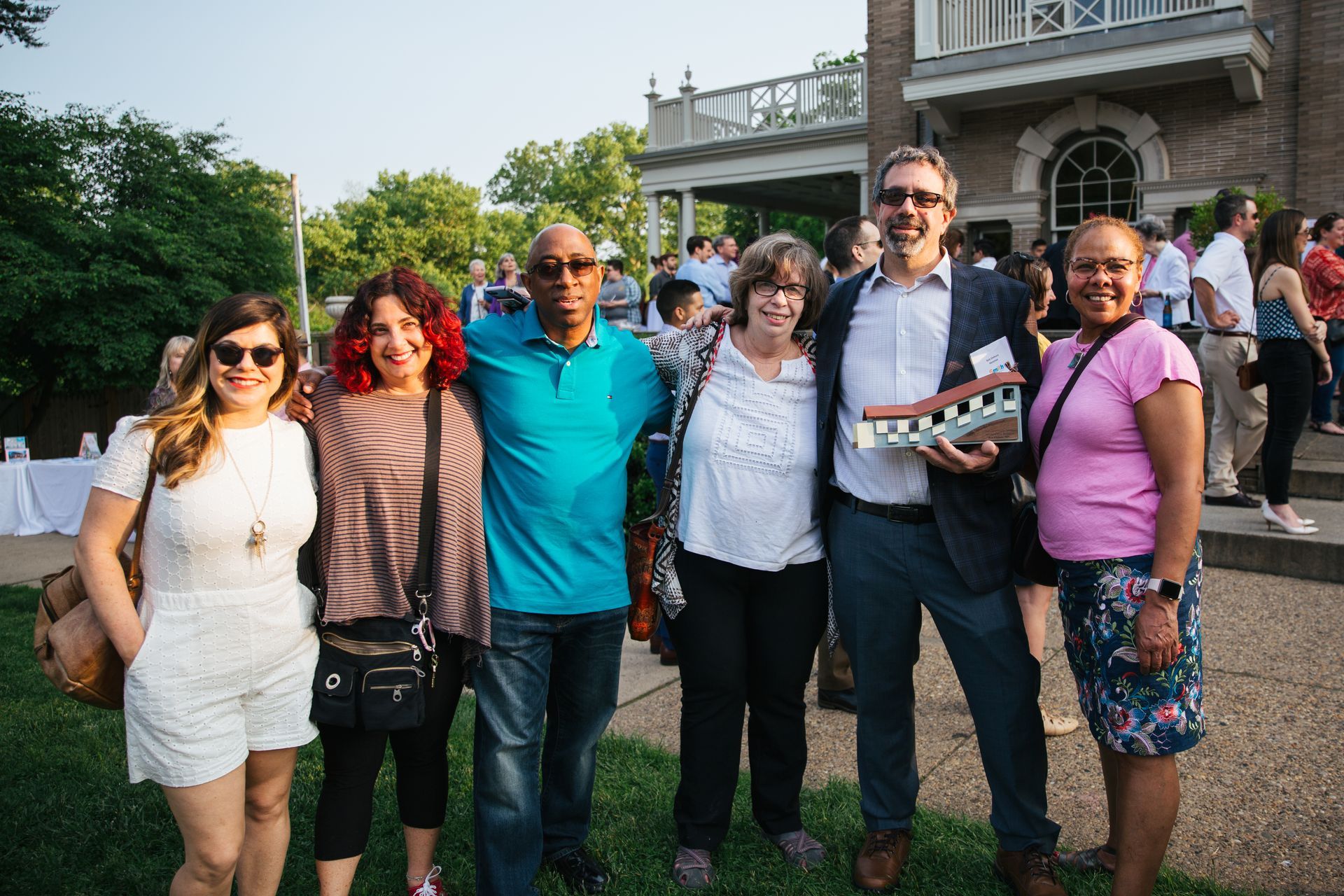 Group of six people posing outdoors on a lawn, smiling in front of a building.