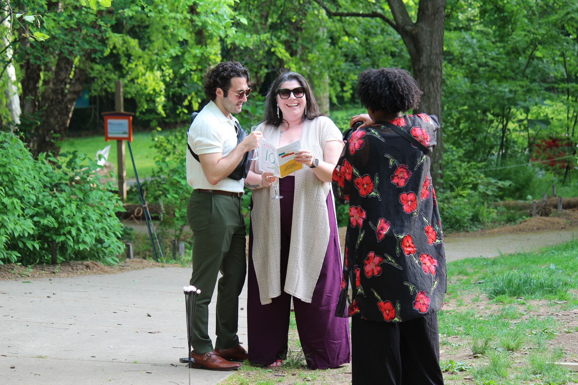 Three people chatting outdoors in a park, holding drinks near a tree-lined path.