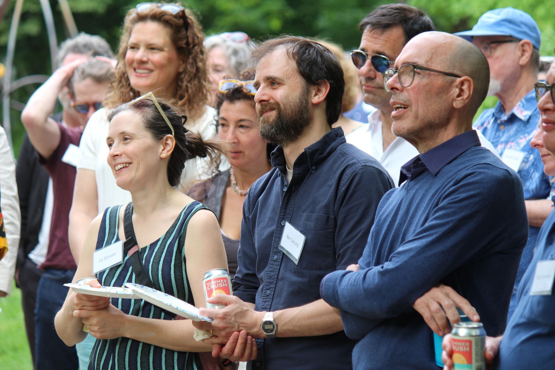 Audience members standing outdoors, smiling and watching an event, one holding a water bottle and papers.