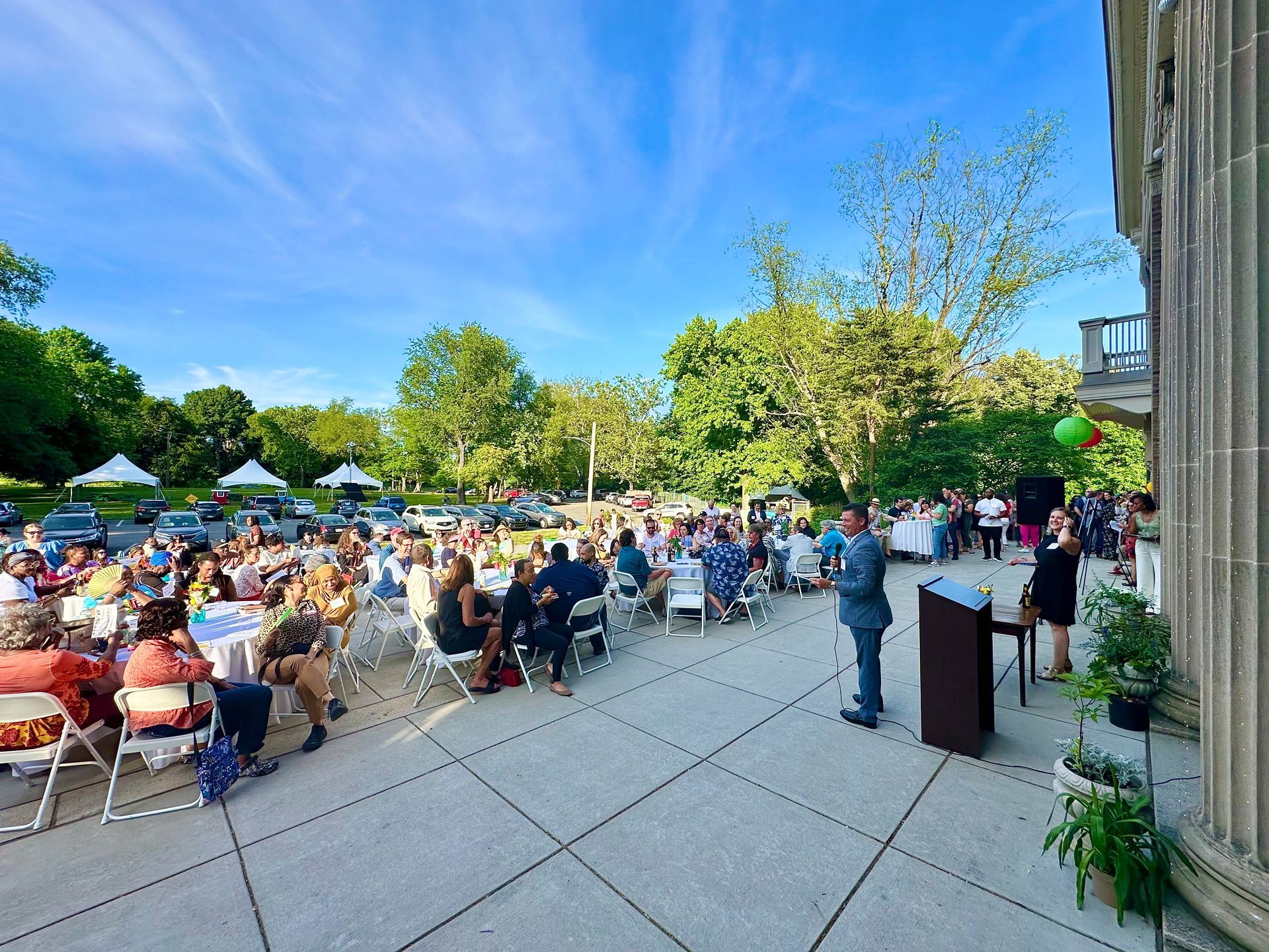 Outdoor gathering with seated crowd on a patio beside a building, speaker at podium under a blue sky