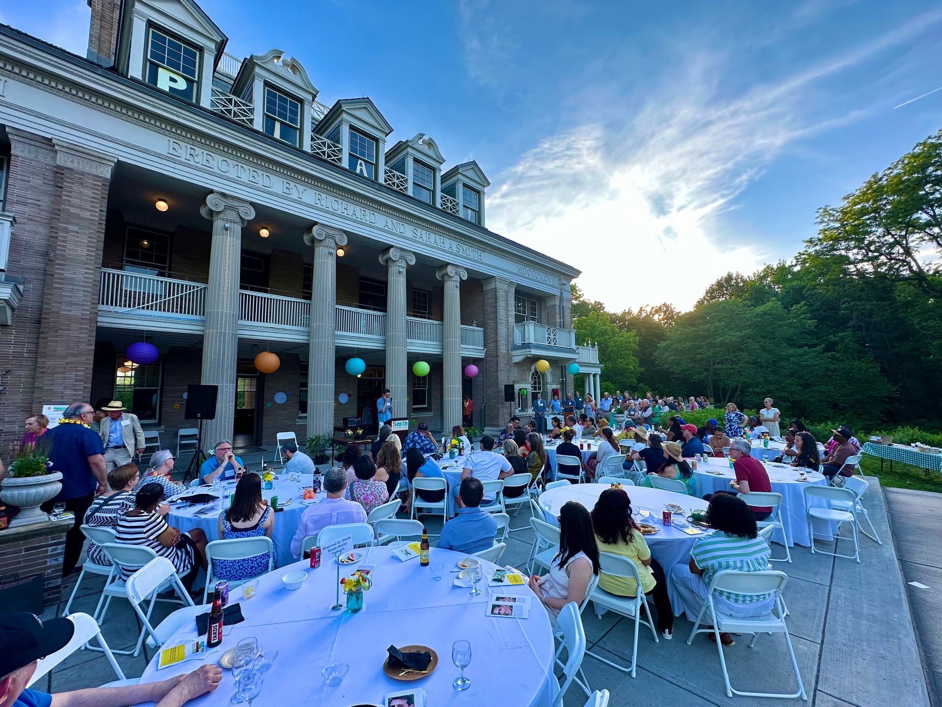 Outdoor banquet with guests seated at long white tables beside a grand historic building at dusk