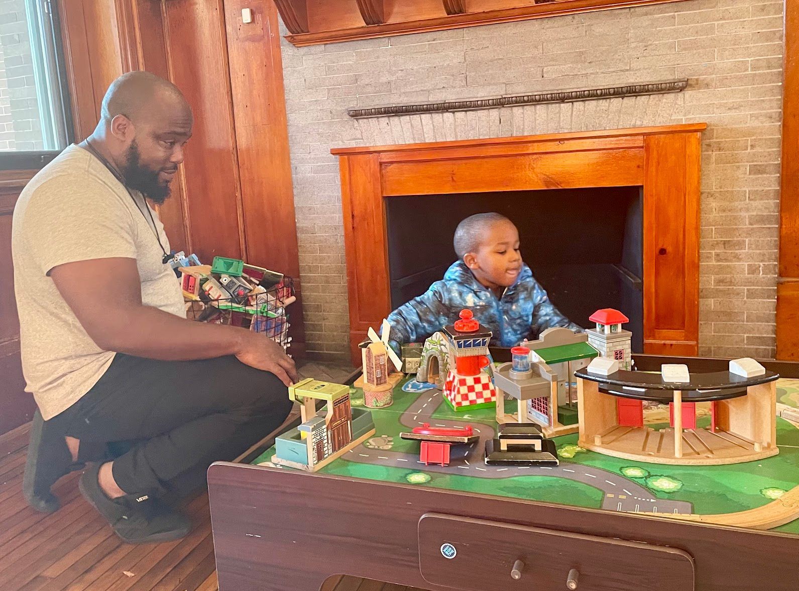 Adult and child playing with a toy village on the floor beside a fireplace