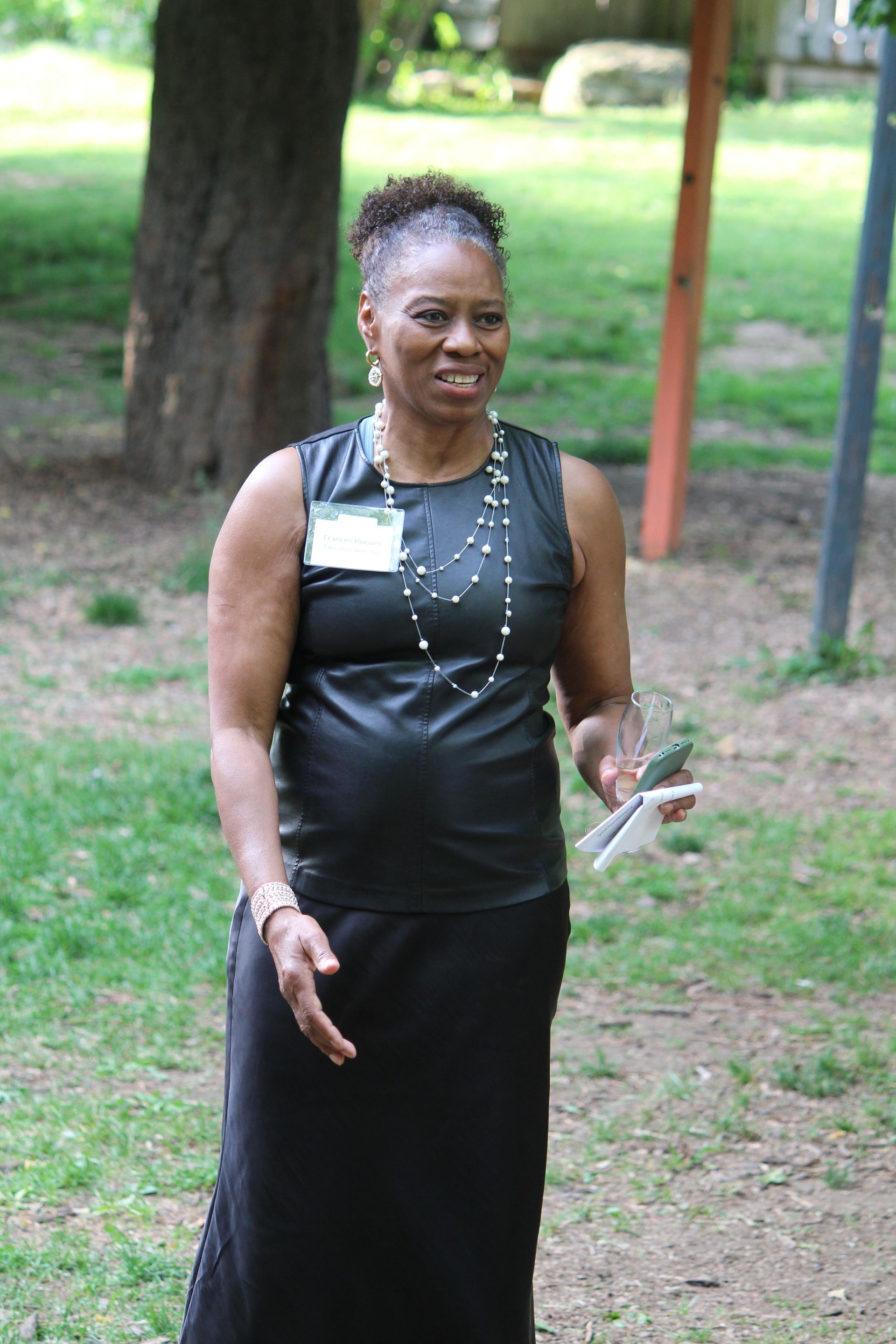 Woman in a black dress standing outdoors in a park, holding flowers and smiling gently.