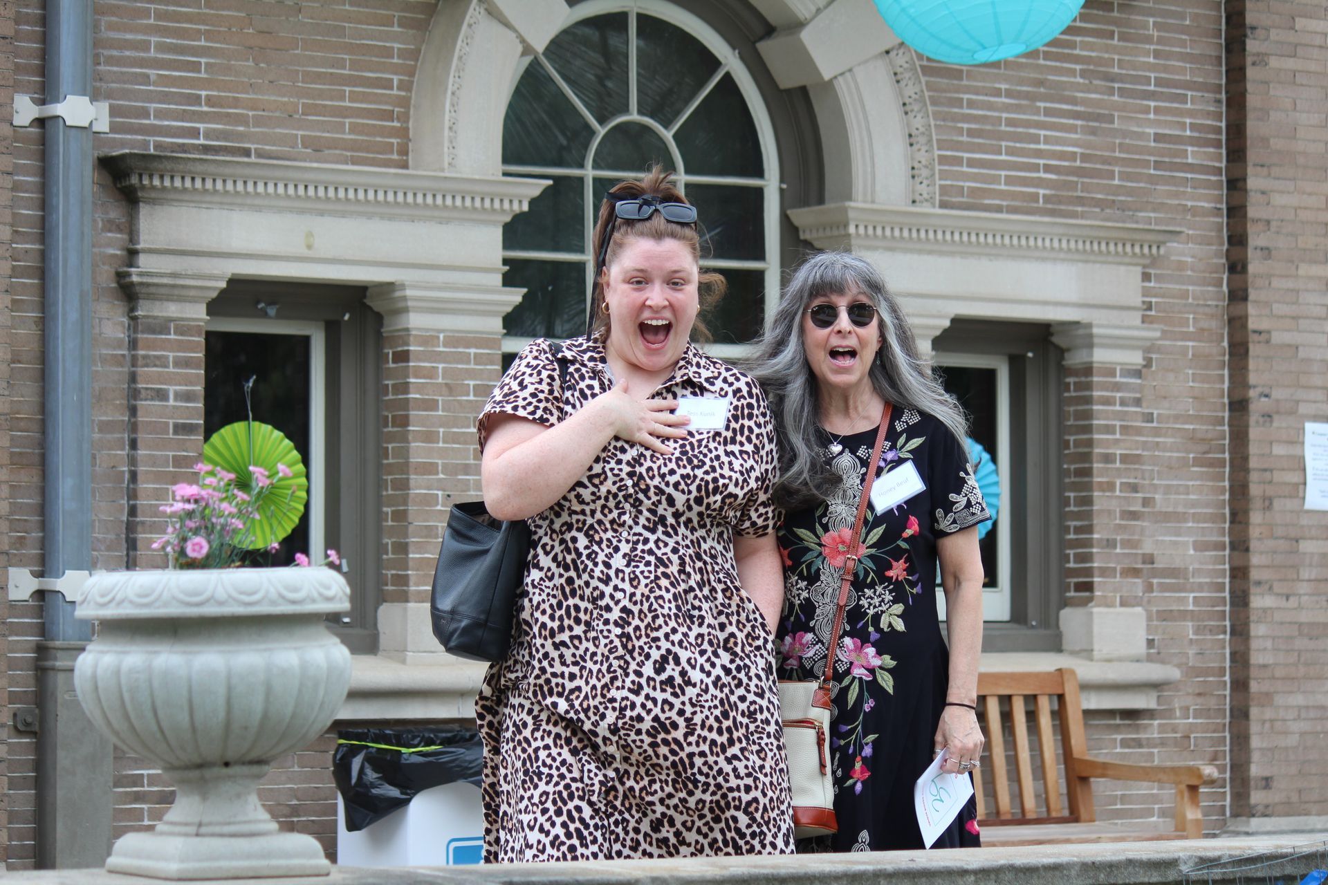 Two women standing on a porch, smiling, with balloons and a brick house behind them.