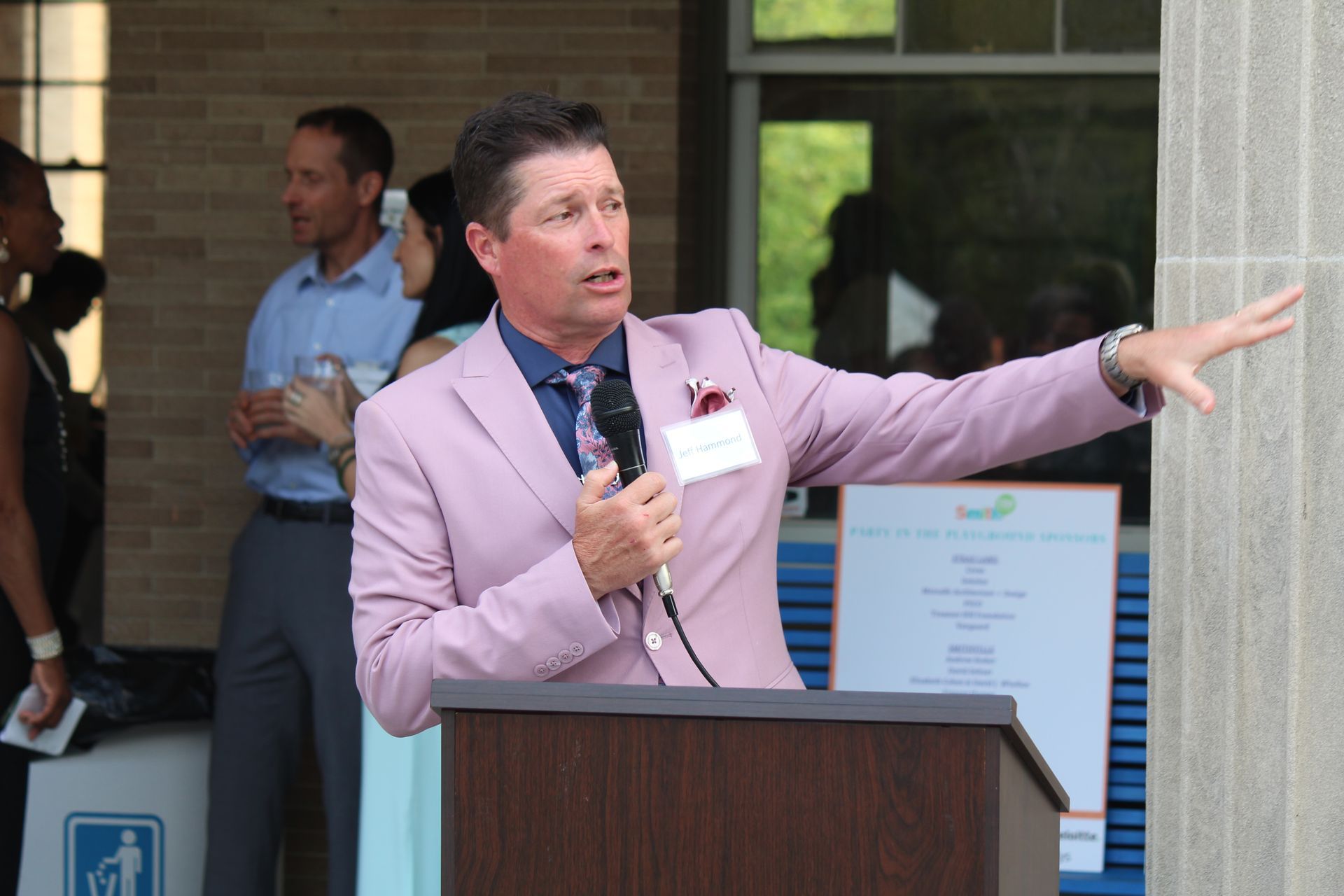Man in a pink blazer speaking at a podium, gesturing outdoors near a building entrance.