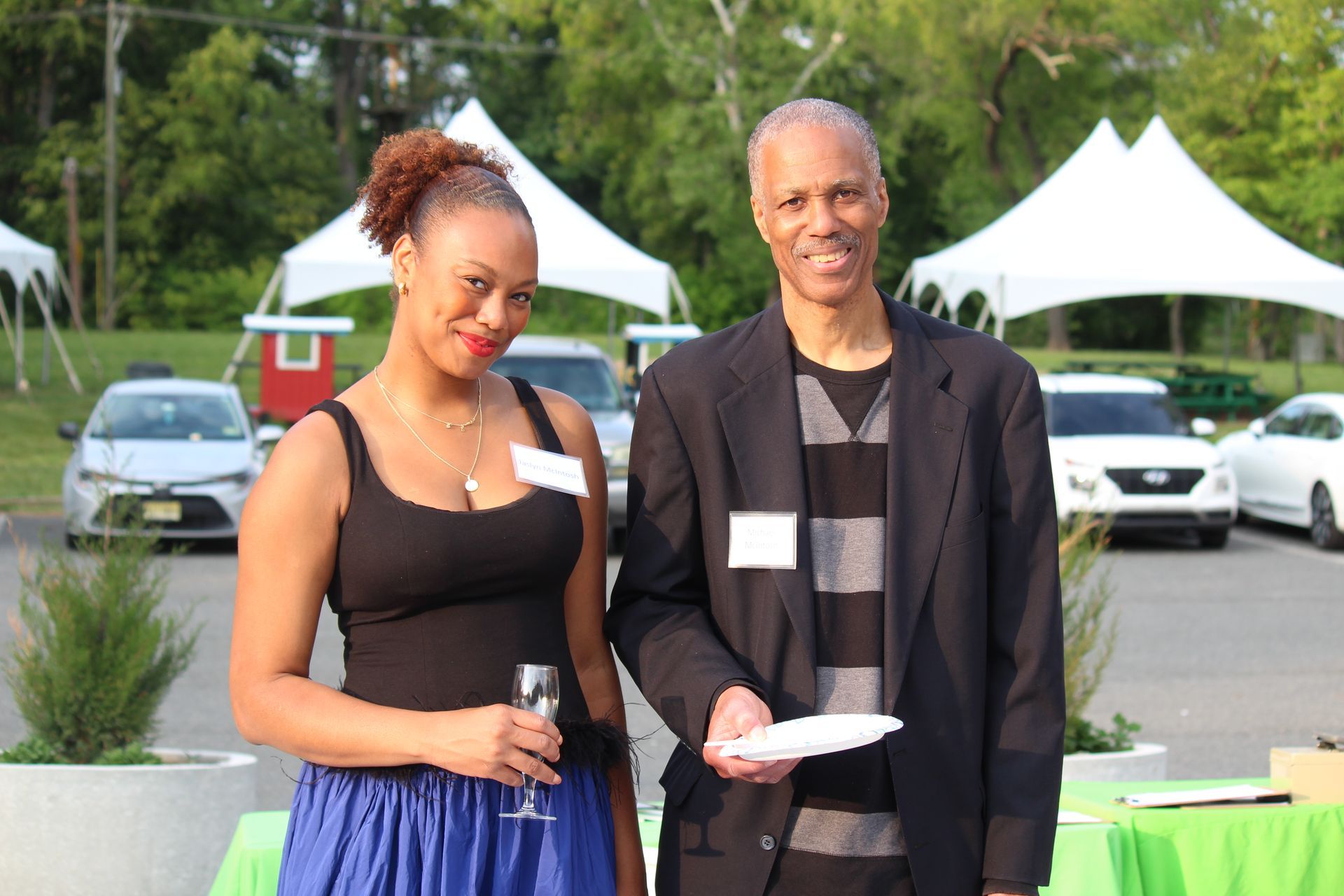 Two people standing outdoors at an event, holding microphones and papers, with cars and tents behind them.
