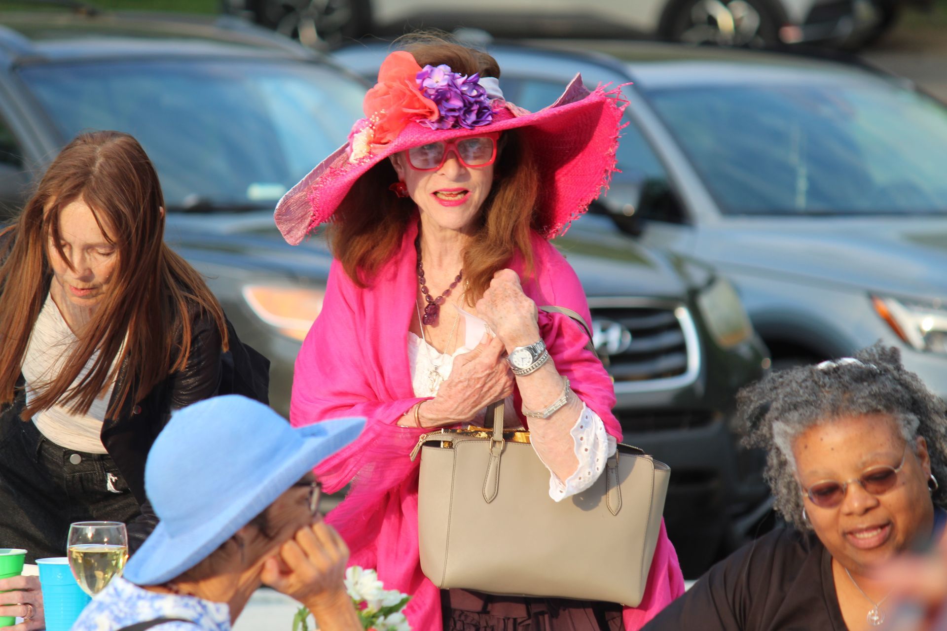 Woman in a bright pink hat and shawl standing at an outdoor event, holding a beige handbag.