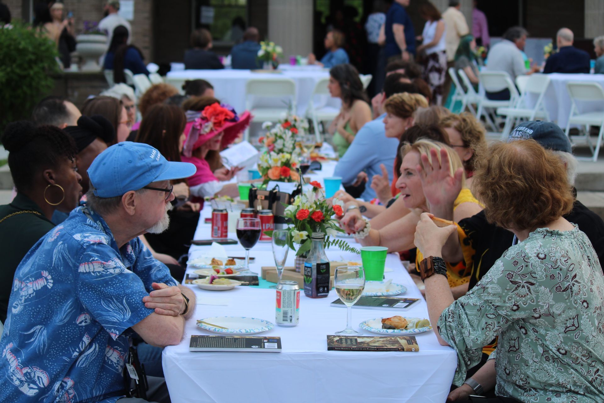 People seated at a long outdoor dinner table with wine, flowers, and conversation at dusk