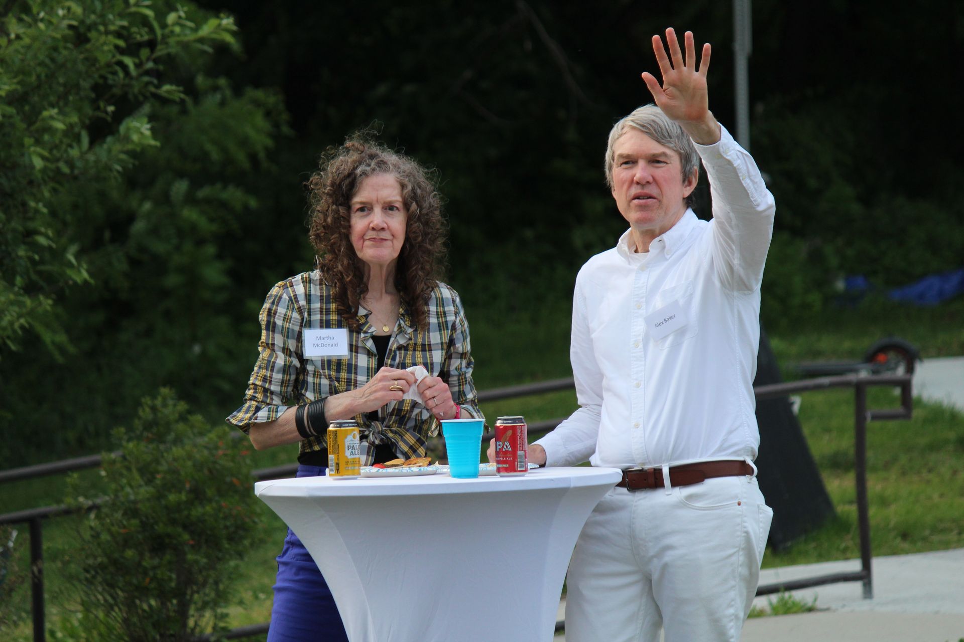 Two people standing at an outdoor table, one waving, with drinks and a dark green backdrop