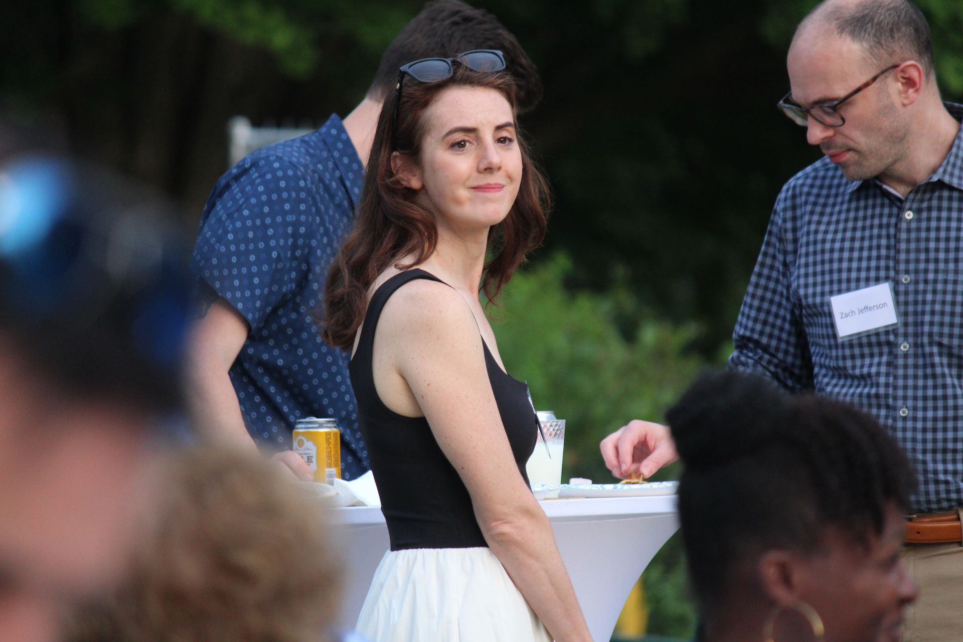 Woman in a black top and white skirt stands at an outdoor gathering, looking toward the camera.