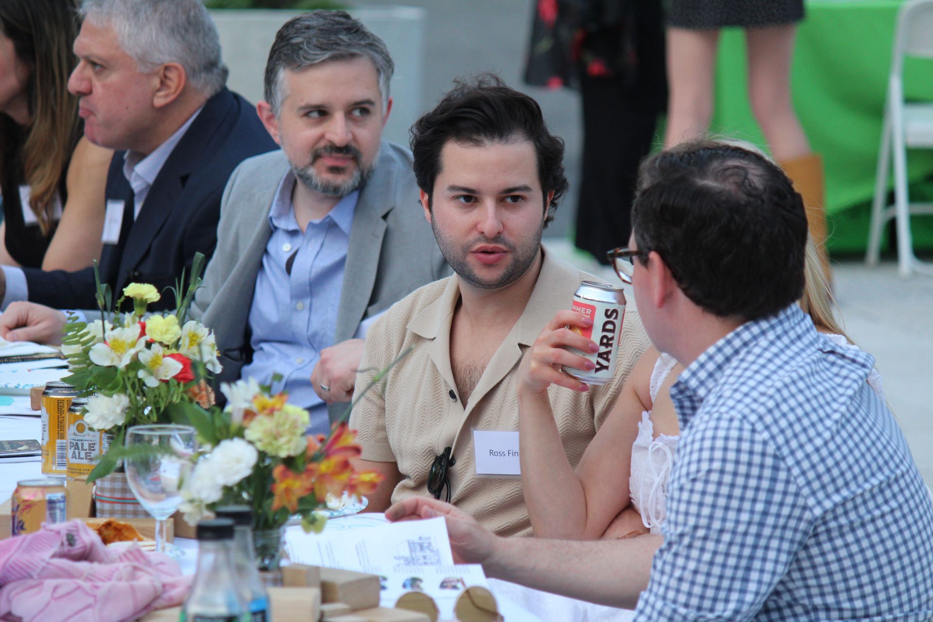 People seated at a banquet table, talking and eating with floral centerpieces and drinks.