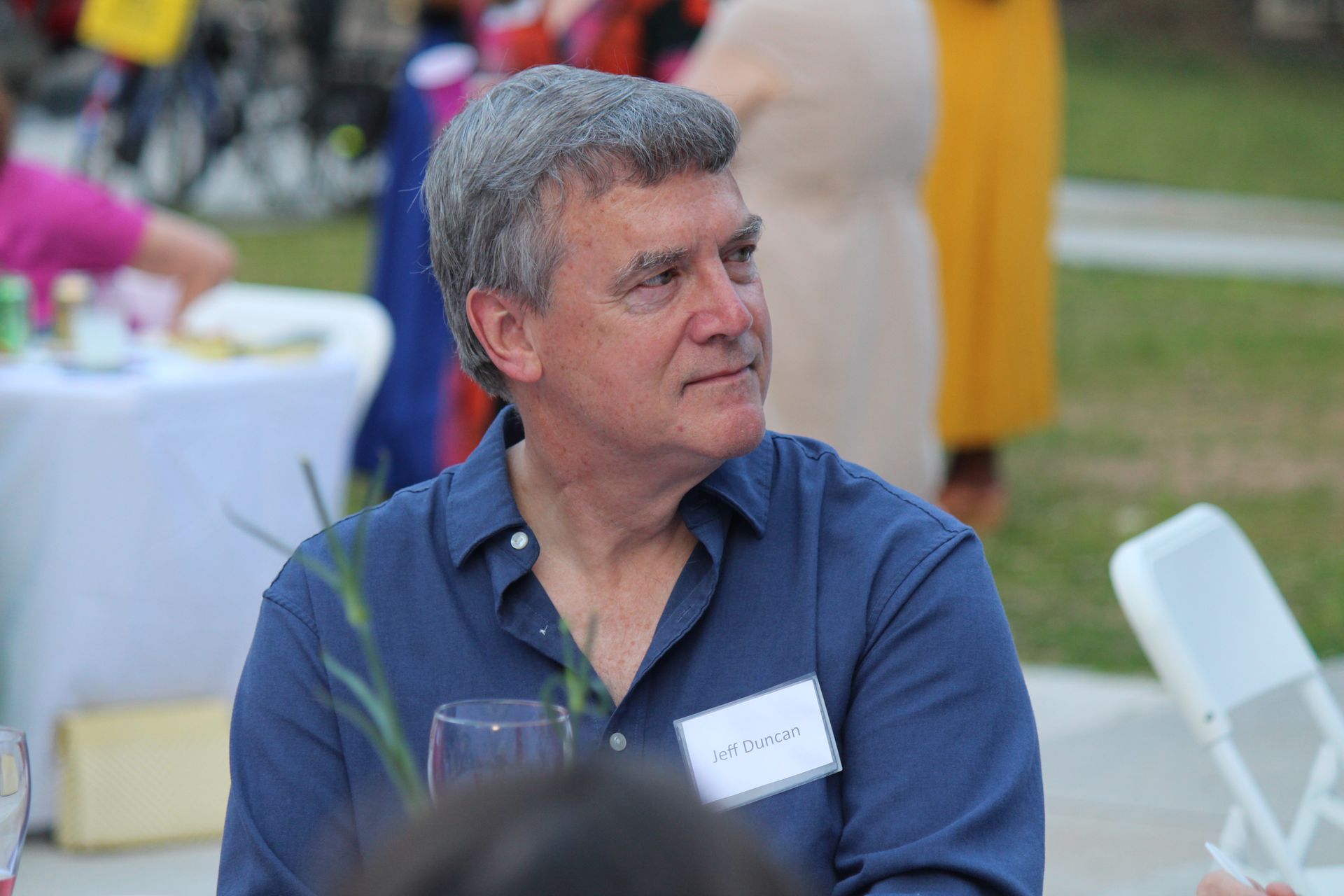 Man in a blue shirt seated outdoors at a table, looking to the side at a social gathering.