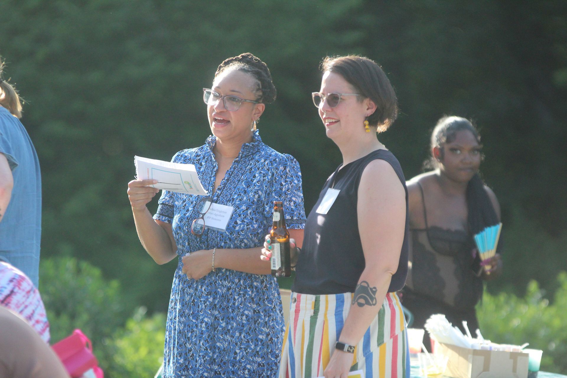 Two women talking outdoors, one holding a drink and the other reading from papers.