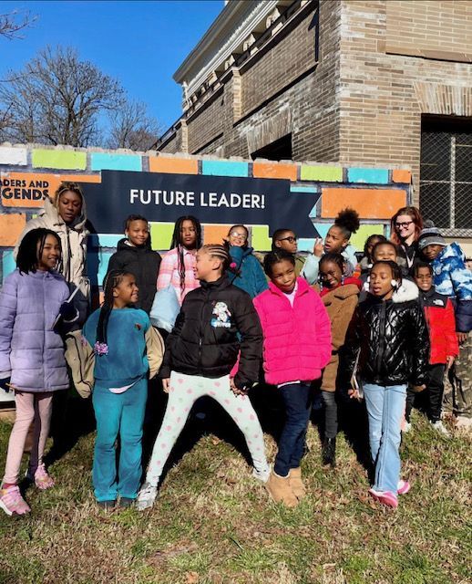 Group of children posing outdoors in front of a “FUTURE LEADERS” banner at a brick building.