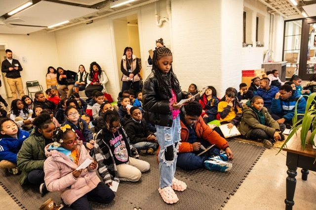 Children seated on the floor in a classroom, listening to a speaker standing at the front.