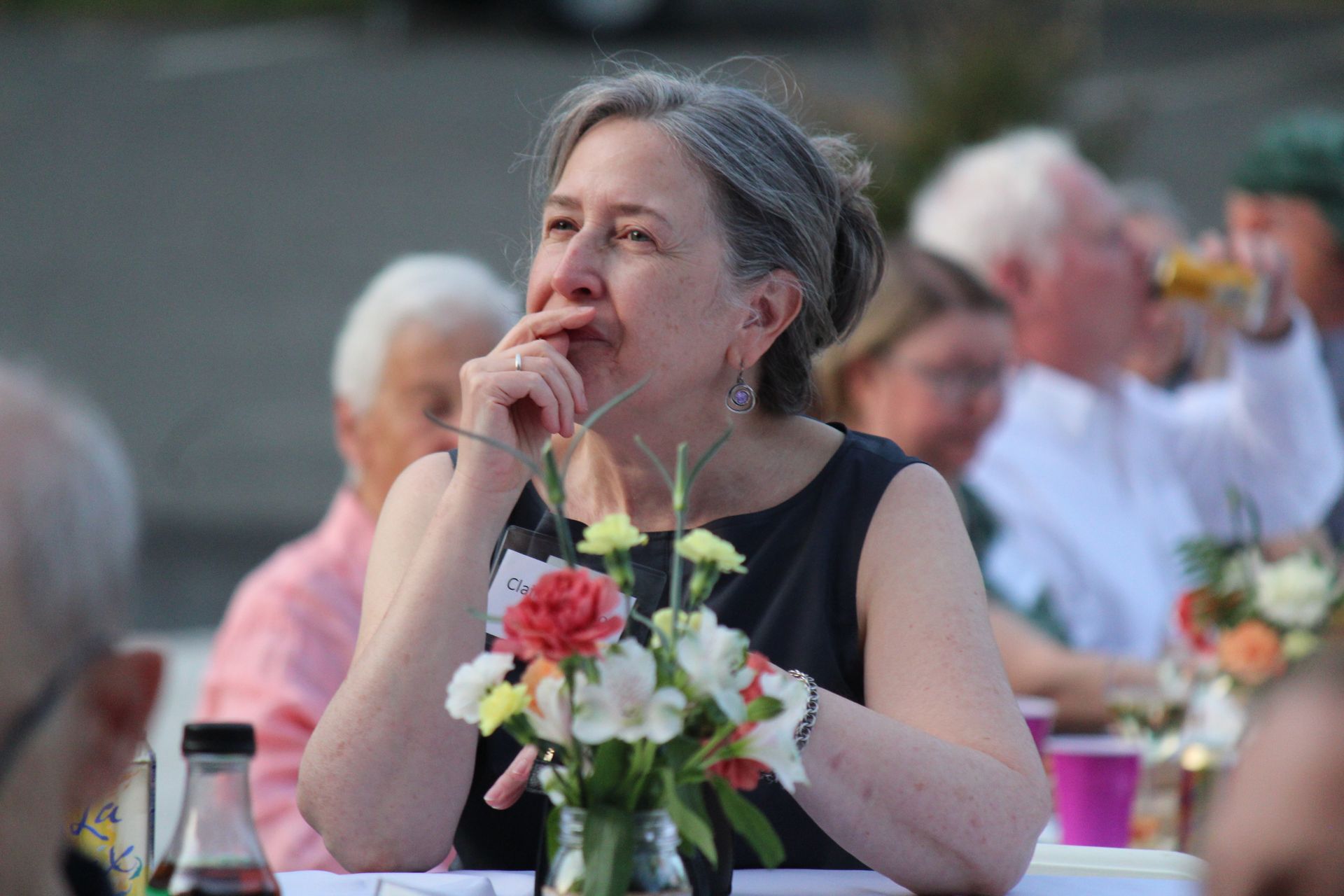 Woman in a black dress sits at an outdoor table, resting her hand on her chin beside a flower centerpiece.