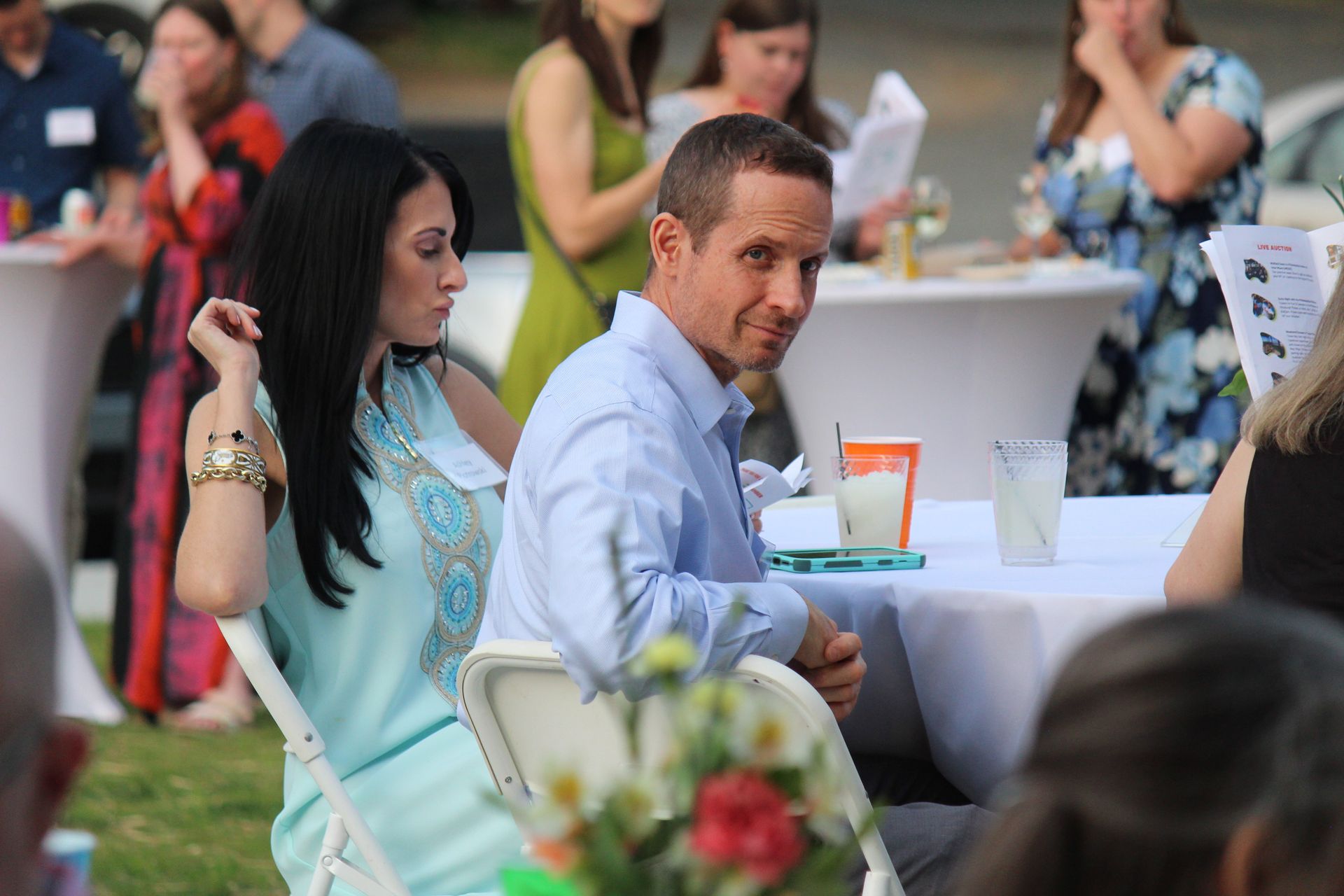 Two people seated at an outdoor event table, with drinks and flowers in the foreground.