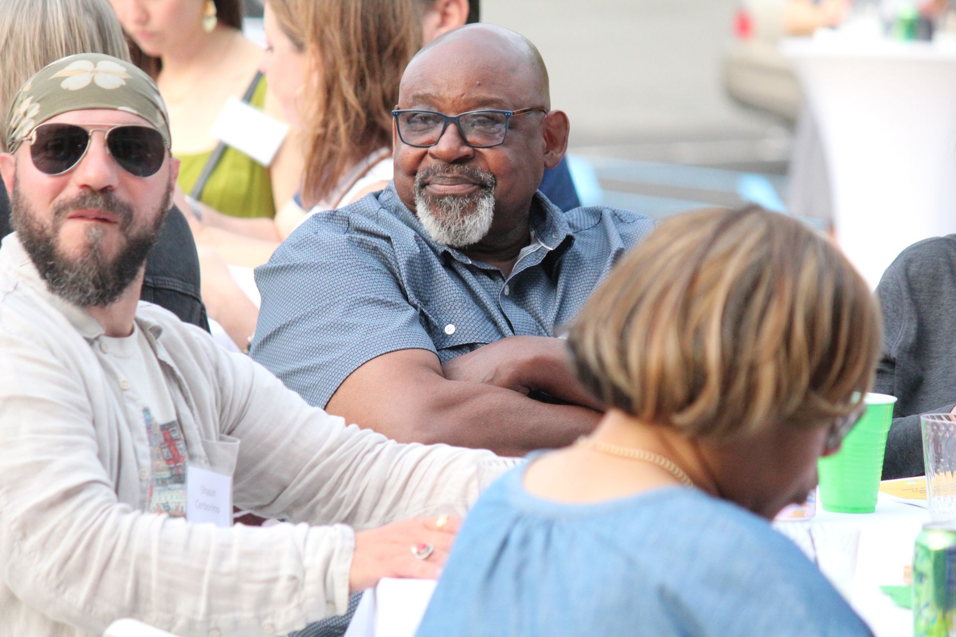 People seated at an outdoor table, including a man in sunglasses and a bearded man, in a bright social setting.