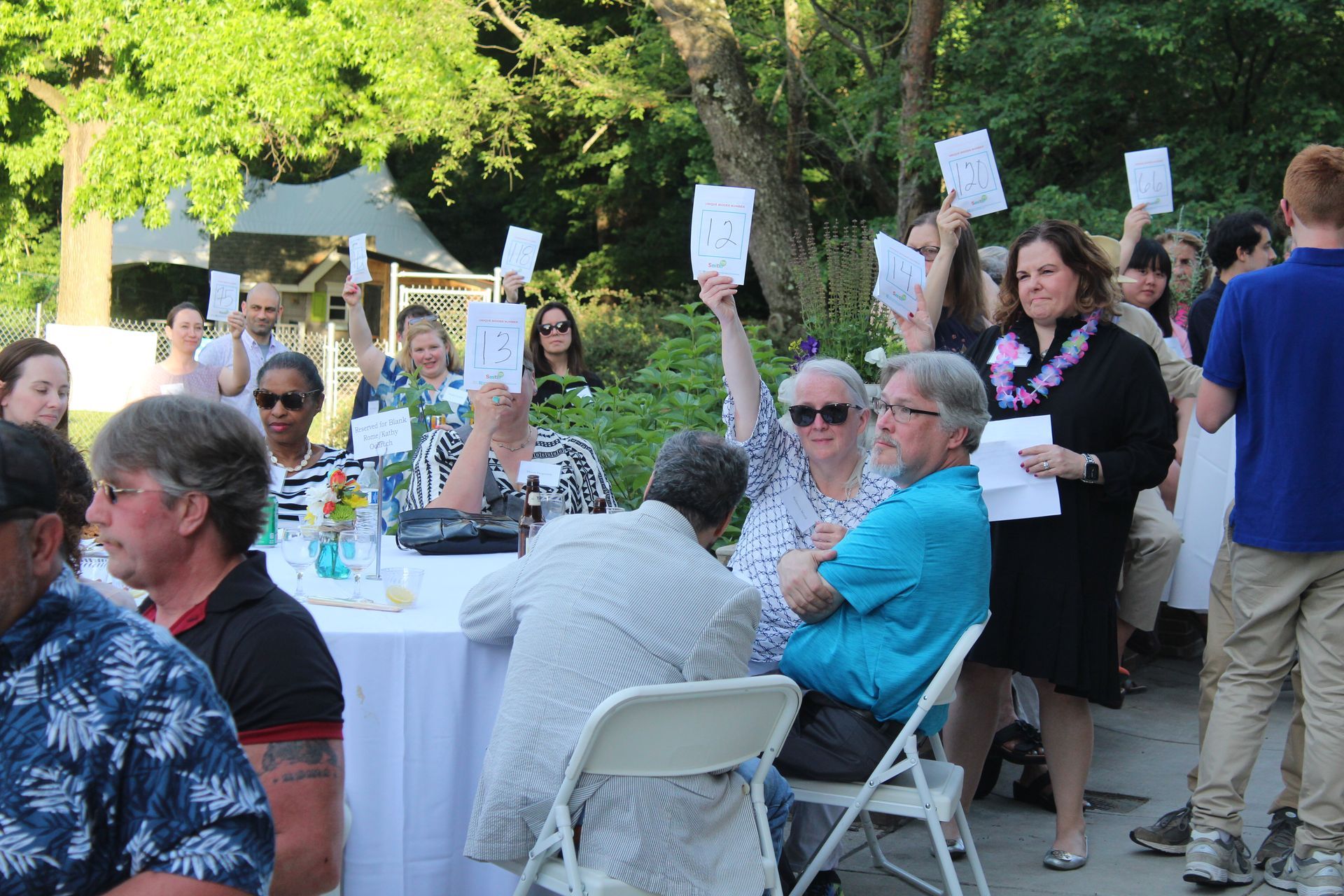 Outdoor gathering at a long table, with seated and standing guests holding signs in a leafy park.