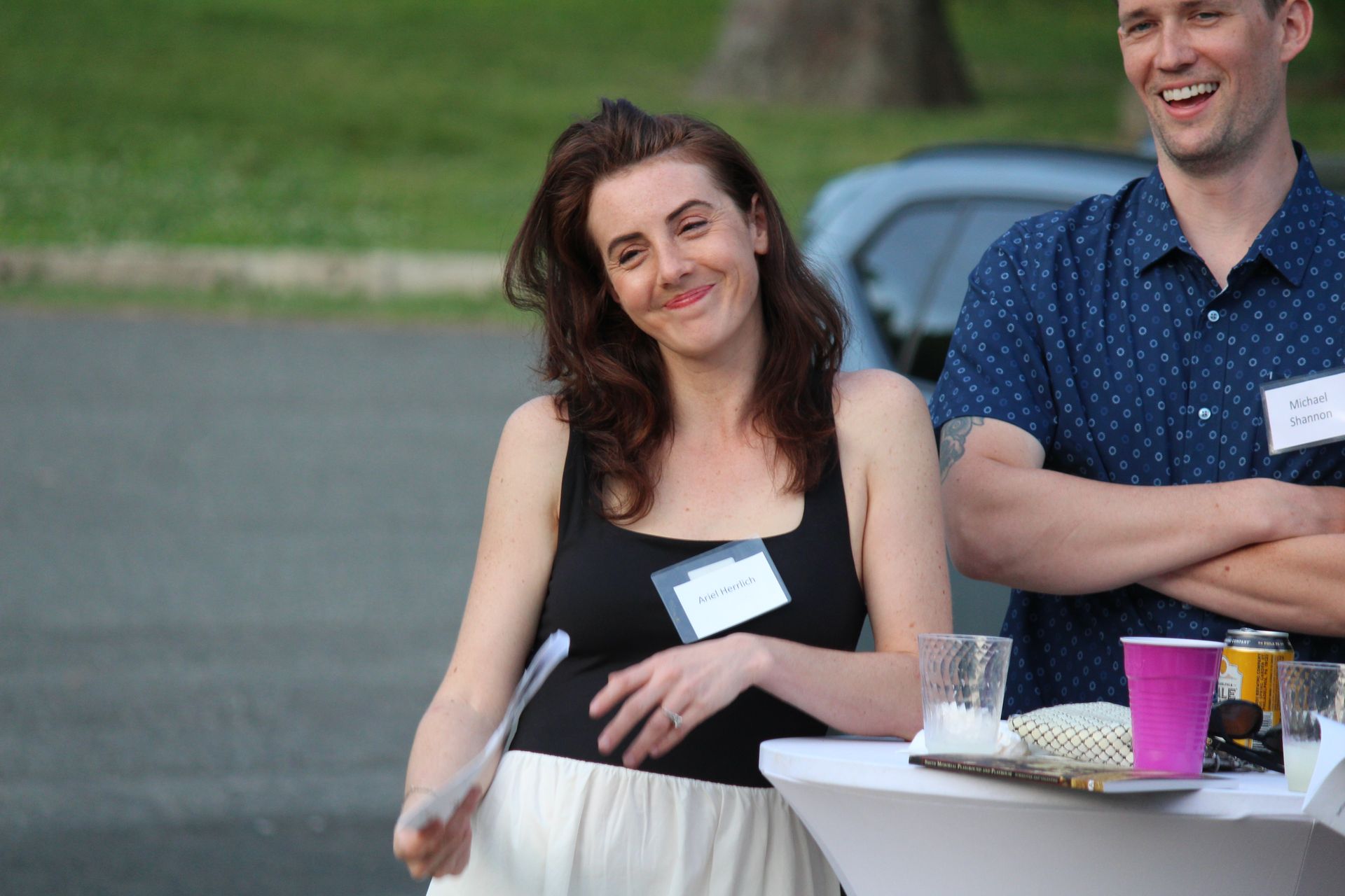 Smiling woman in black top at an outdoor gathering, standing beside a man near a table with drinks and cups