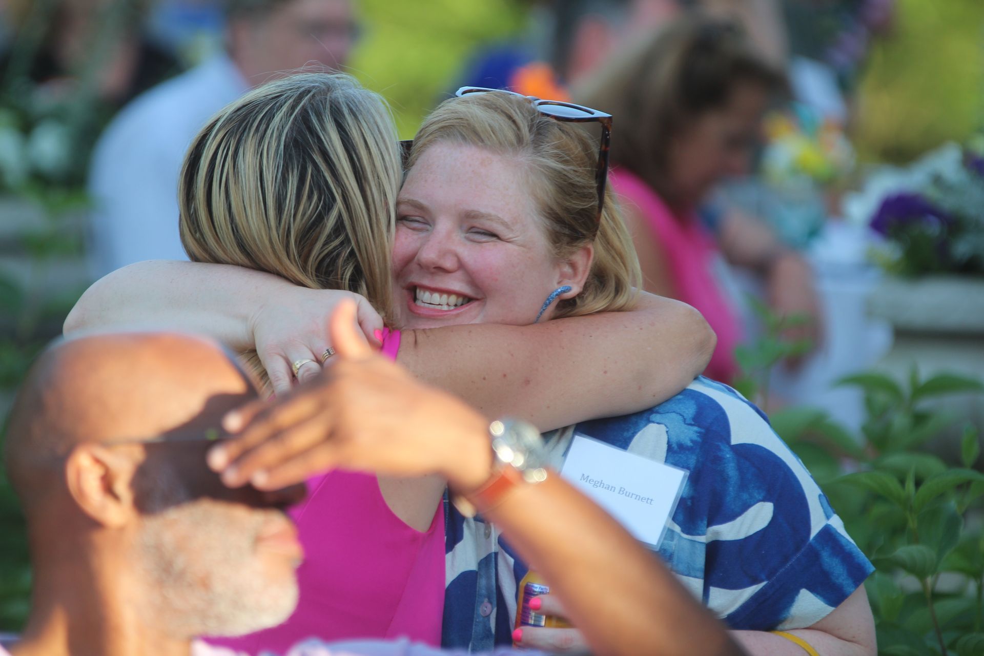 Two people hugging and smiling outdoors, with a blurred crowd in the background.