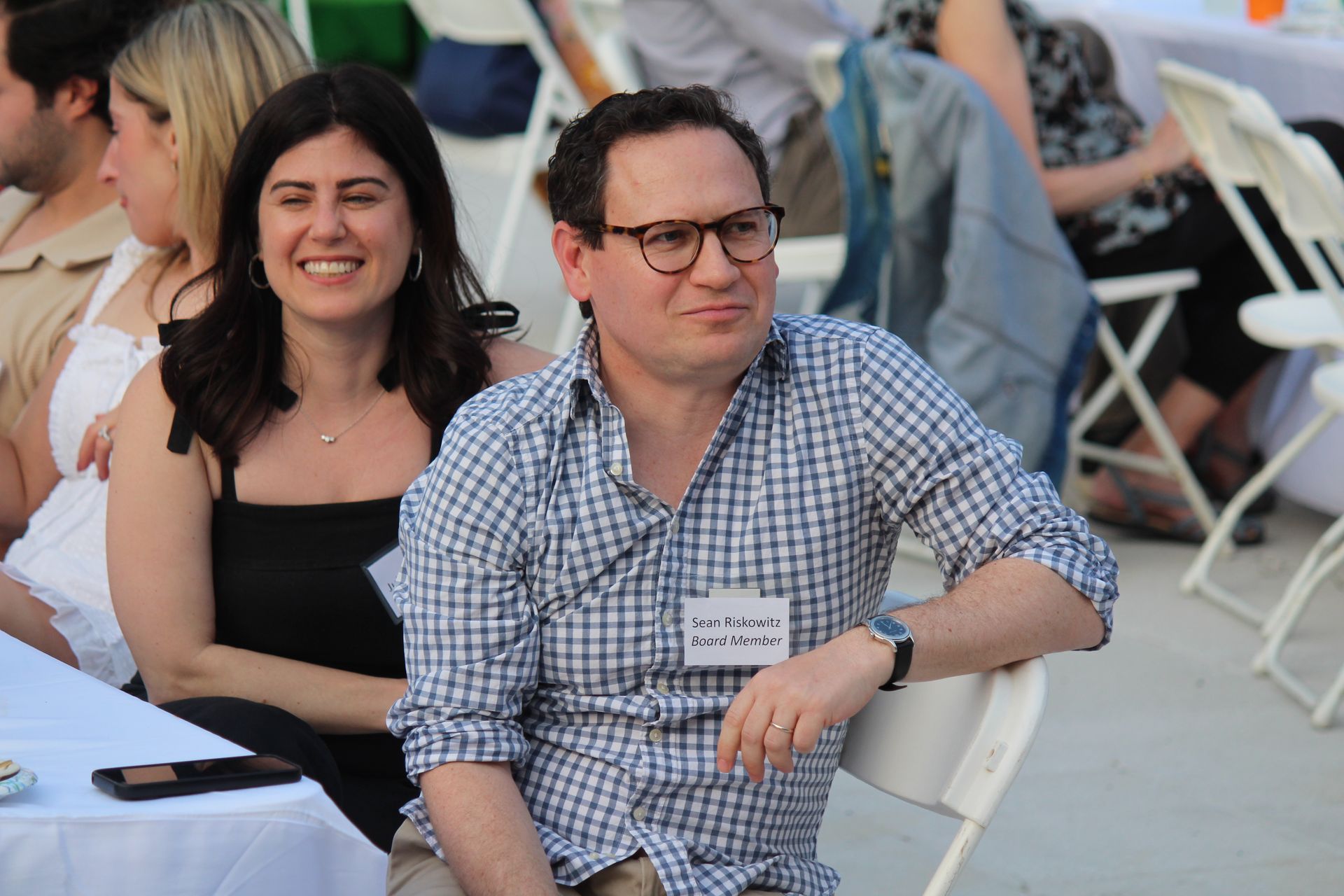 Two seated people at an outdoor event, smiling and wearing casual shirts.