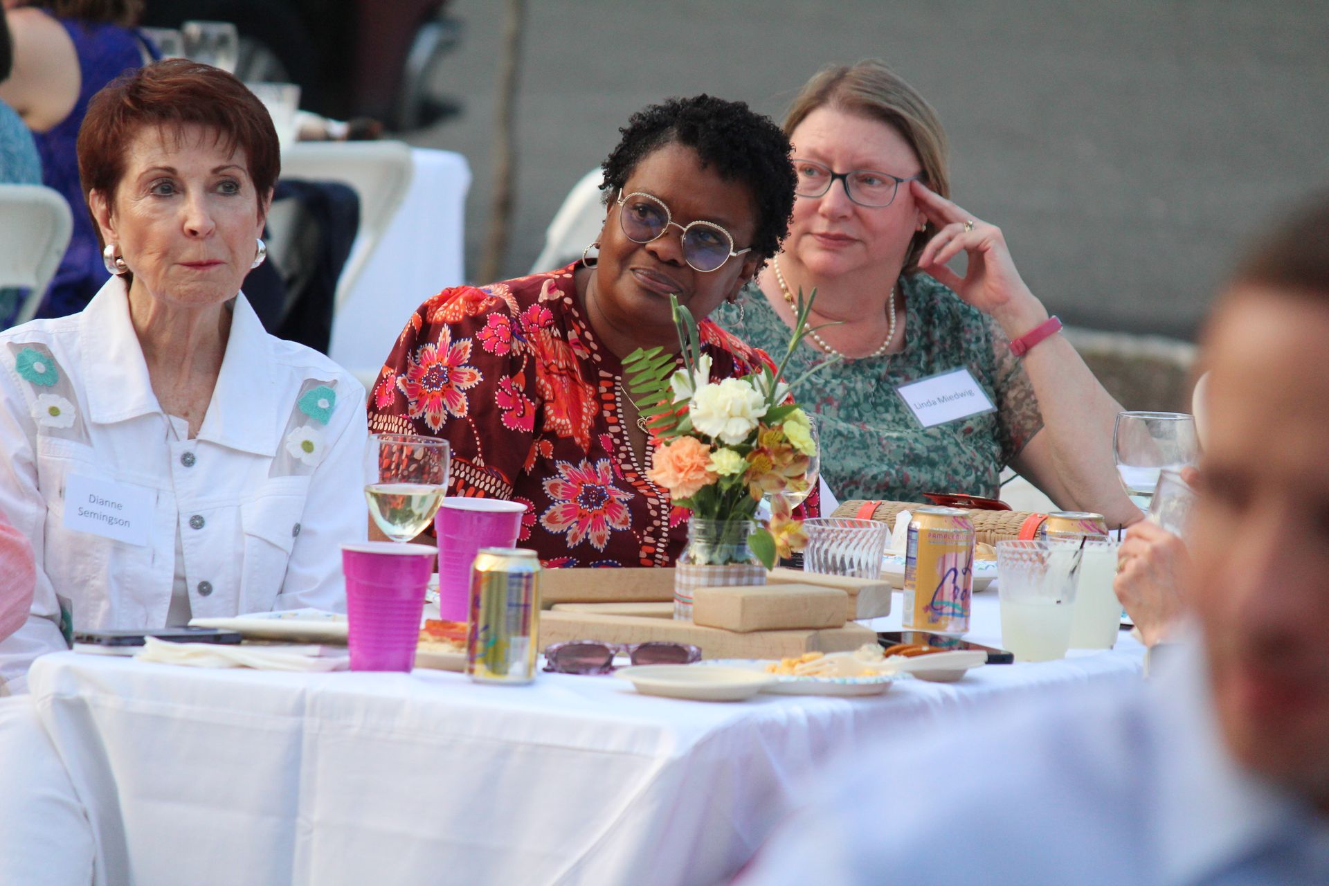 People seated at a banquet table, with drinks, flowers, and plates in a bright outdoor setting