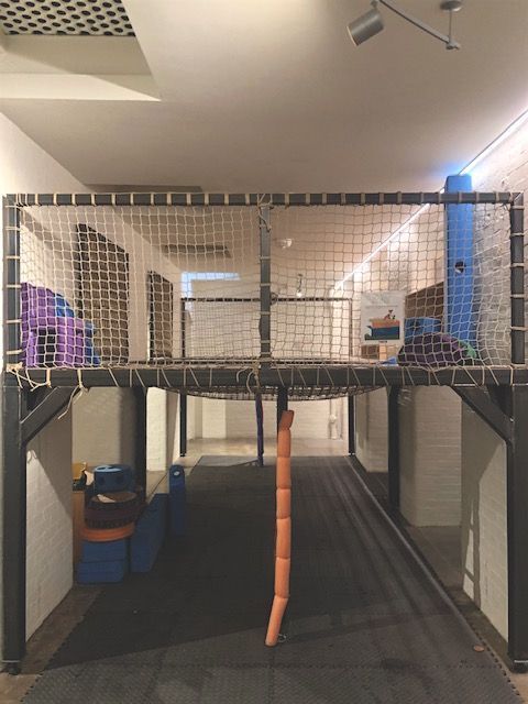 Indoor play area with a raised netted play structure, orange hanging steps, and blue supports.