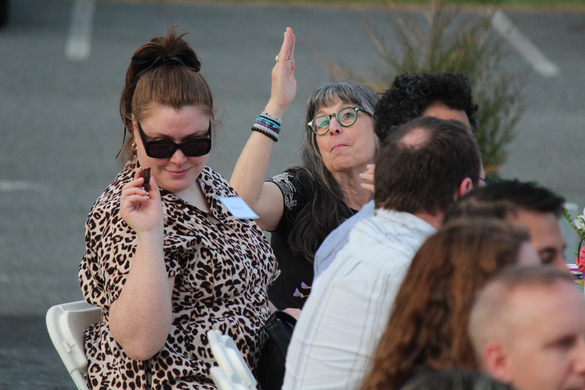 People seated outdoors, one woman in sunglasses raises her hand while others chat nearby