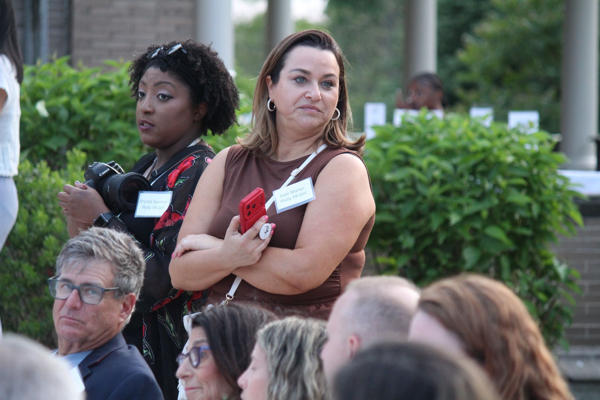 Audience members standing outdoors, one holding a red phone, listening attentively in a garden setting