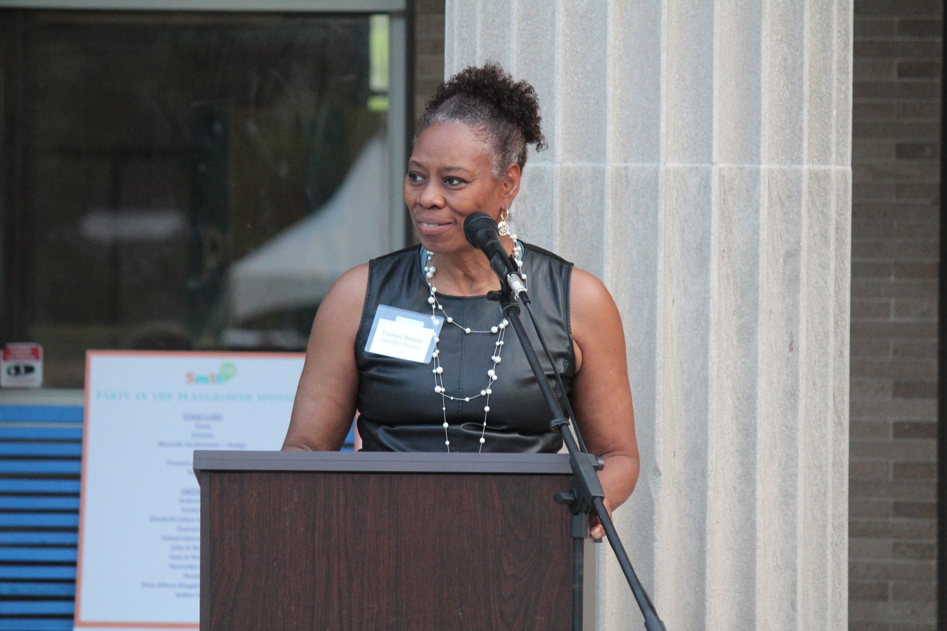 Woman speaking at a podium indoors beside a stone column and microphone.