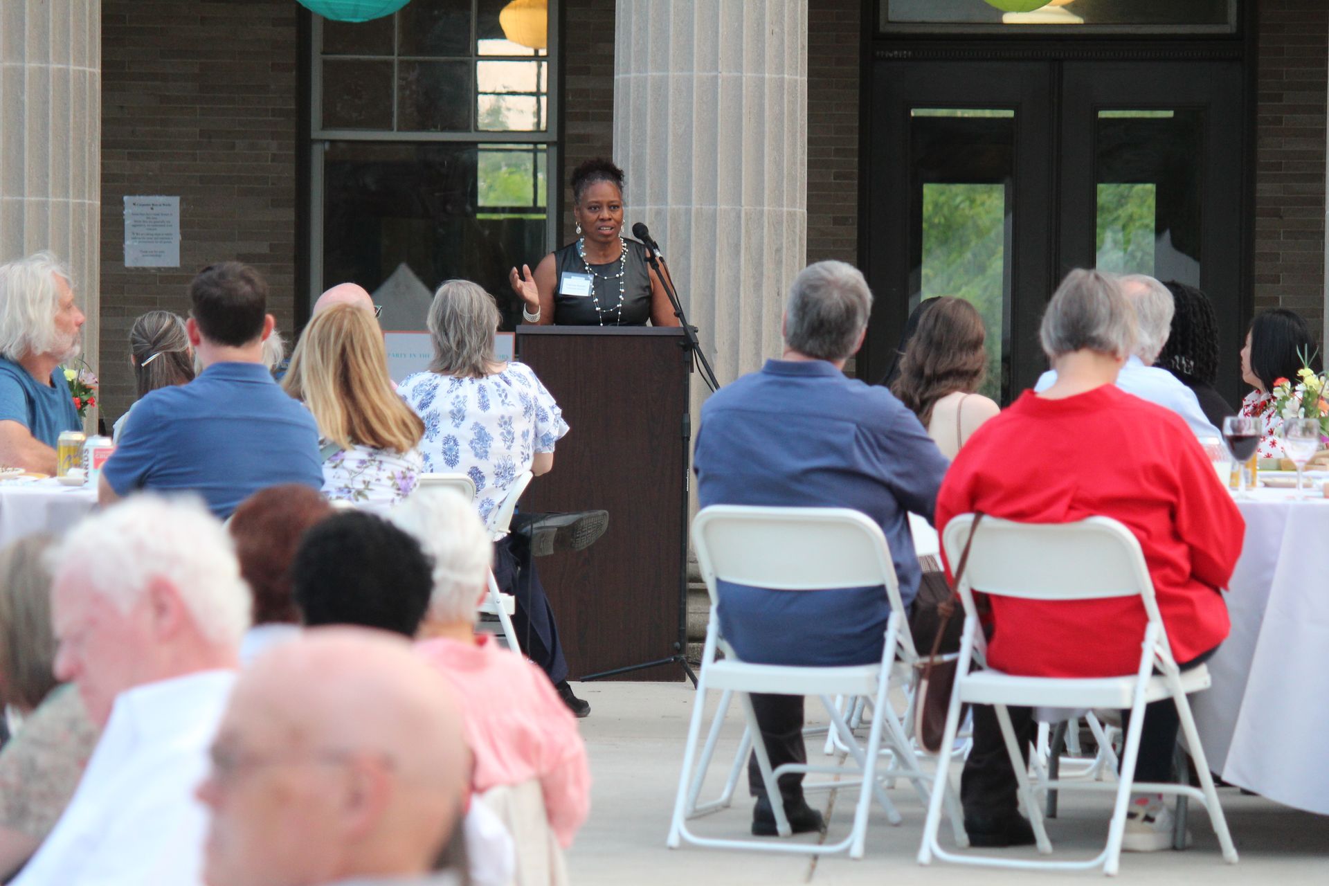 Audience seated outdoors listening to a speaker at a podium in front of a building