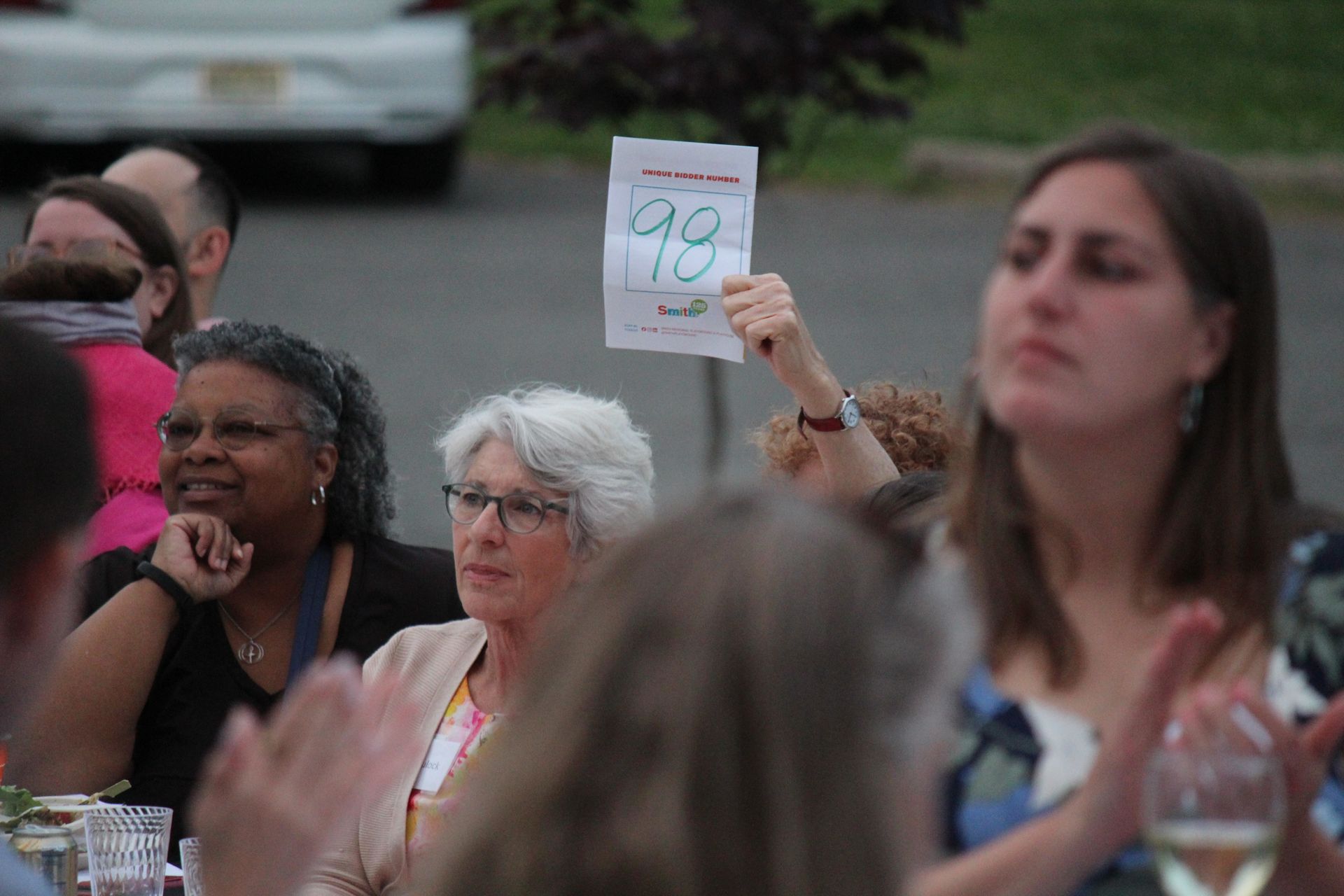 People seated outdoors, one holding a handwritten sign, appearing to applaud at a gathering