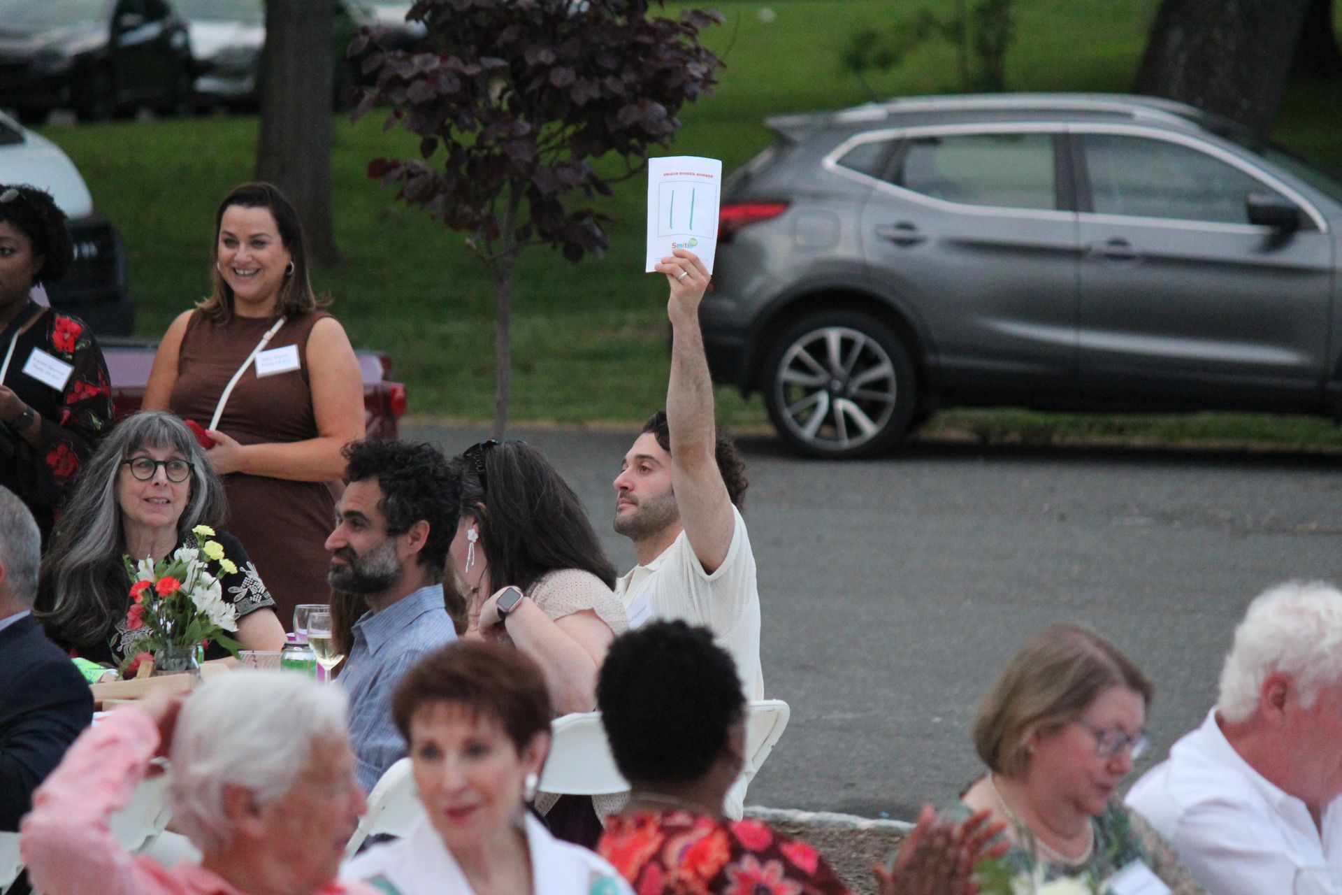 People gathered outdoors, one person raising a white card amid a crowd near parked cars.