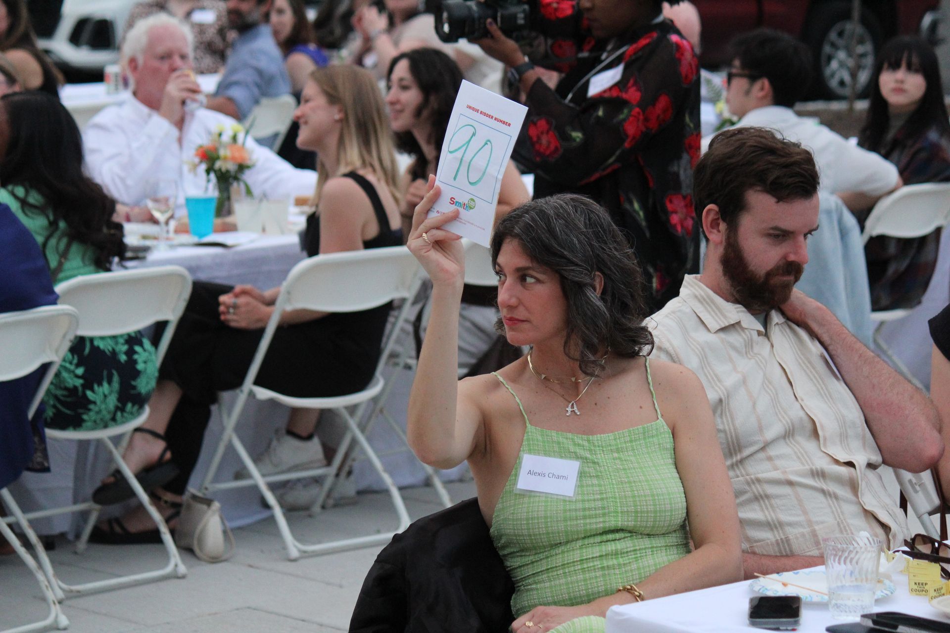 Seated woman in a green dress raises a paper at an outdoor event with tables and chairs