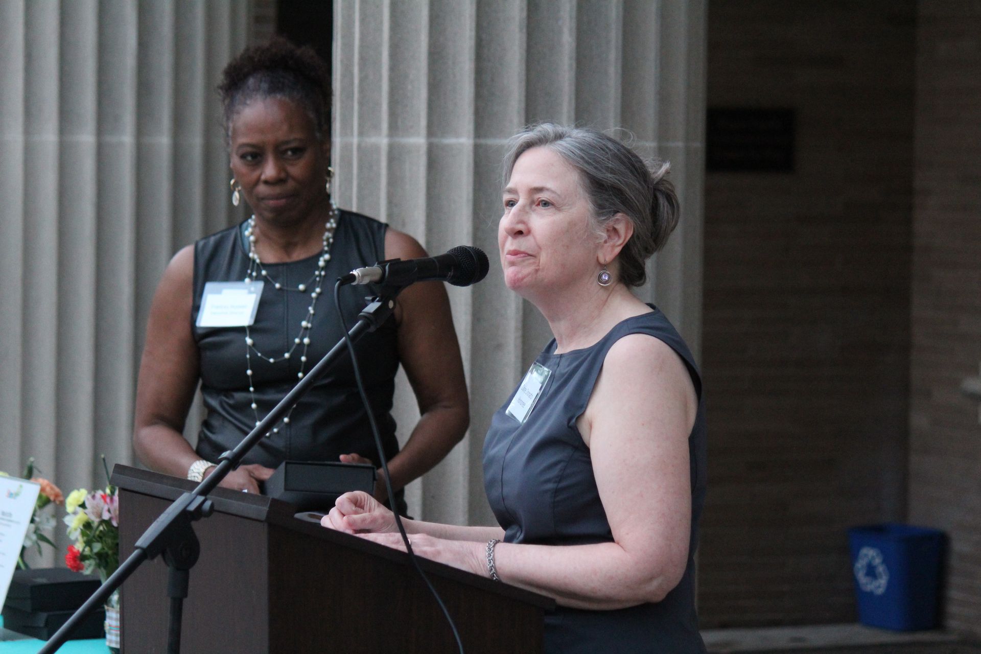 Two women speaking at a podium, one in black and one in gray, with microphones in front of a building.