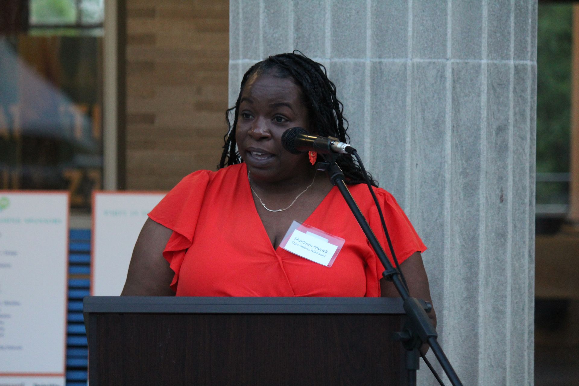 Woman in a red blouse speaking at a podium with a microphone in a columned indoor setting