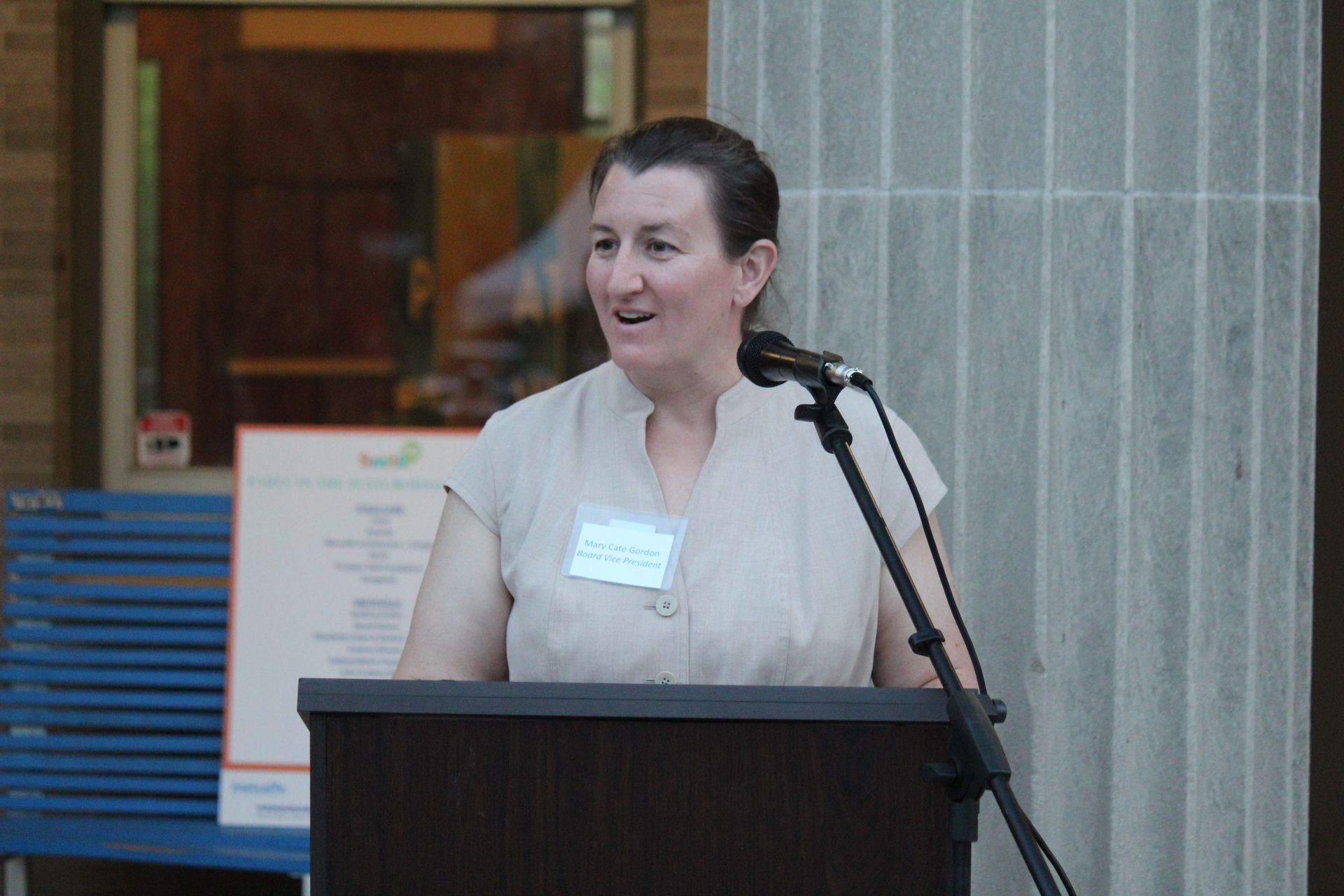 Speaker at a podium with microphone, speaking indoors near a tiled wall