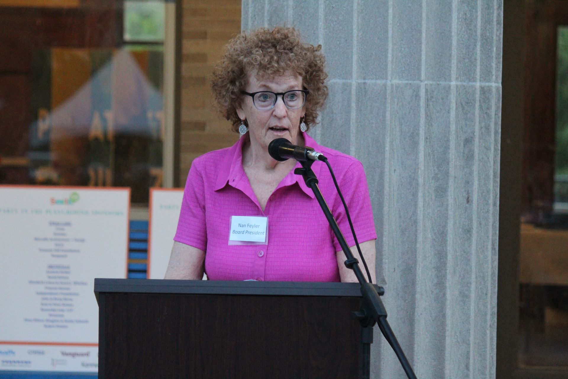 Woman in a pink shirt speaking at a podium with microphones indoors