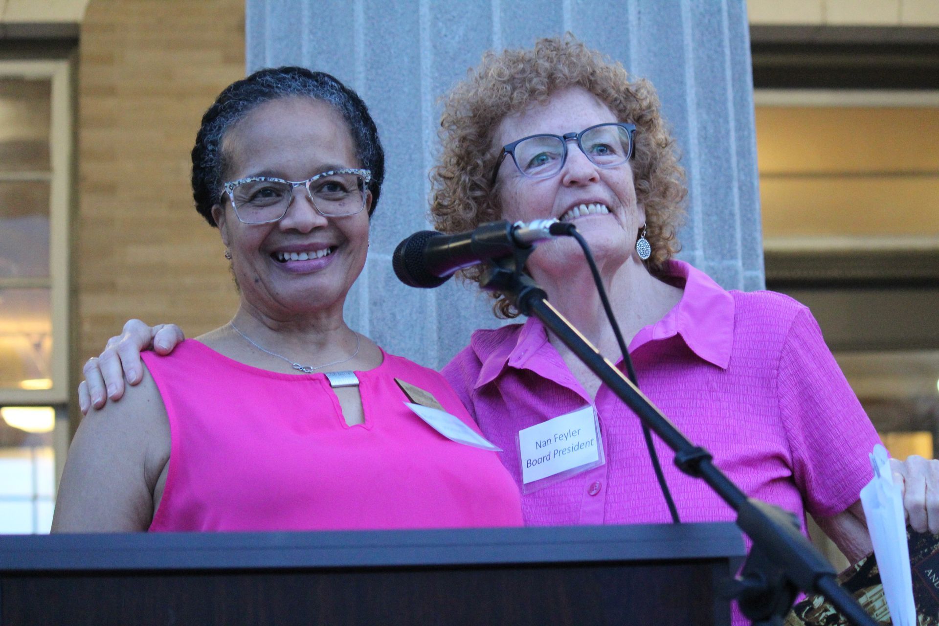 Two women in pink stand at a podium, smiling beside a microphone.