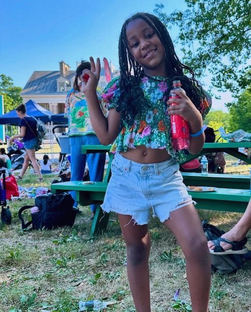 Person smiling and posing outdoors in a floral top and denim shorts at a picnic gathering.