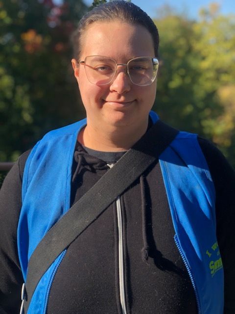 Person wearing a blue vest and glasses, smiling outdoors in sunlight.