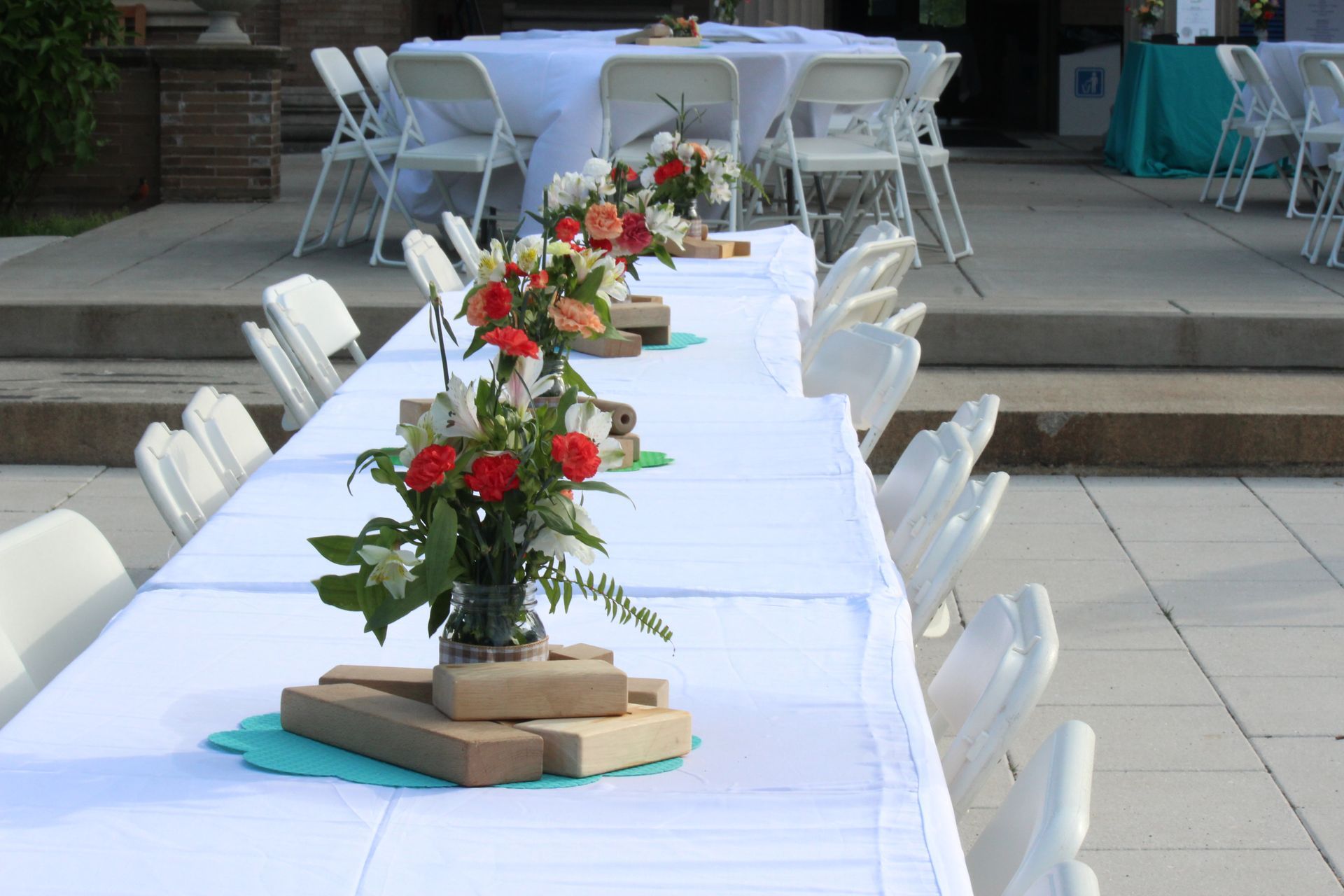 Long outdoor banquet table with white tablecloth, floral centerpieces, and white chairs on a patio.