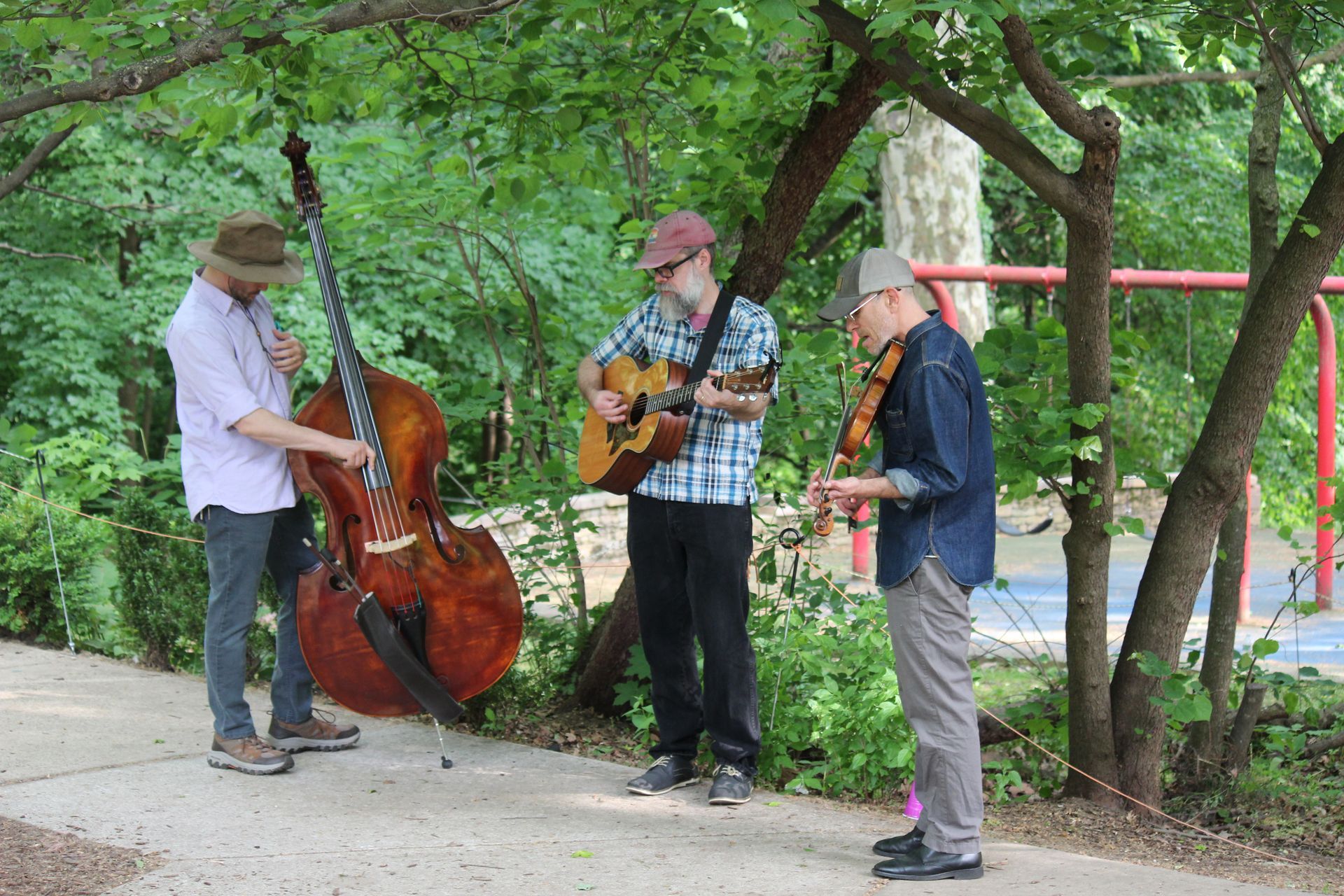 Three musicians play guitar, upright bass, and banjo outdoors under green trees.