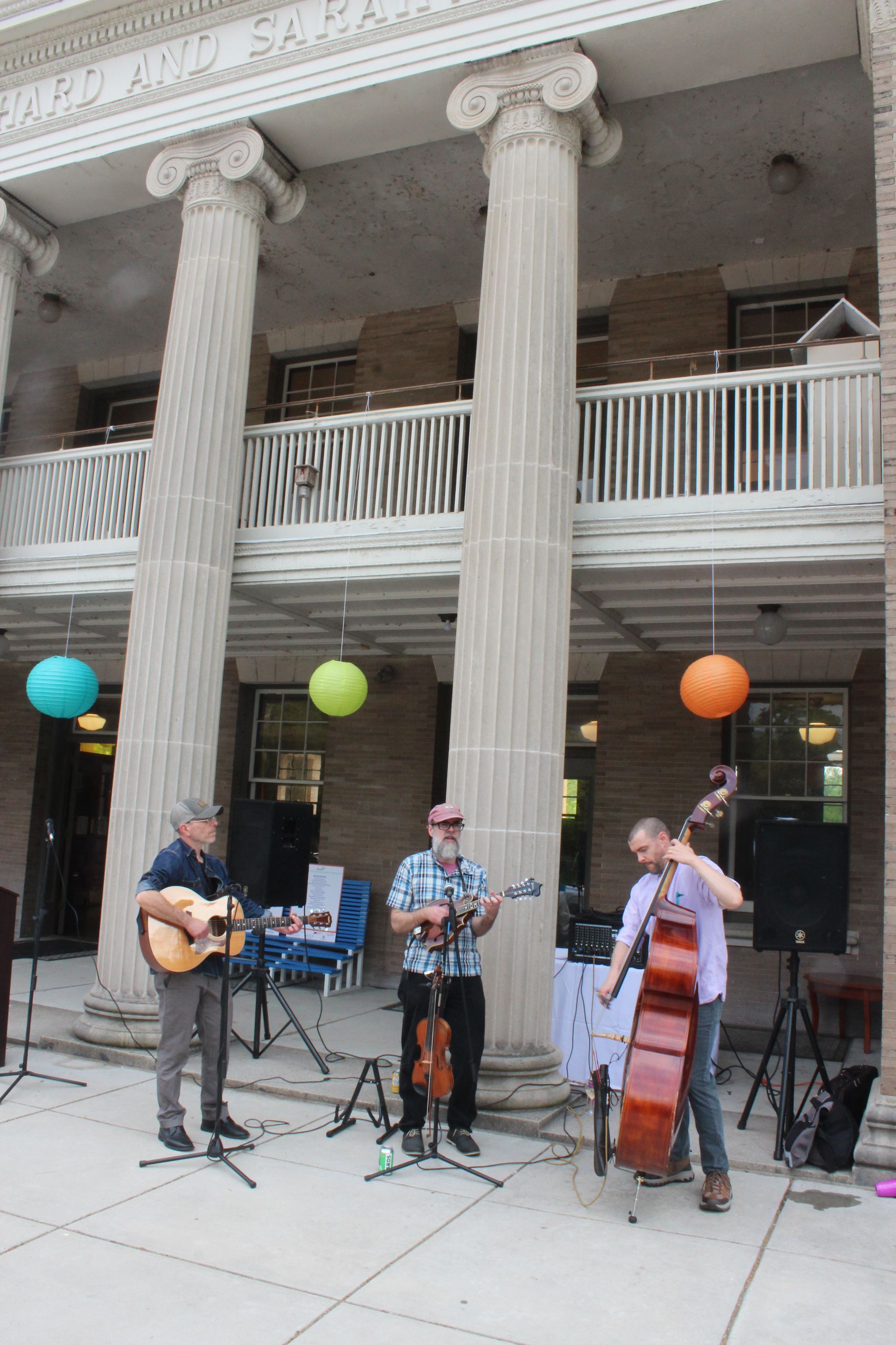 Three musicians perform outdoors before a building with tall columns and colorful balloons.