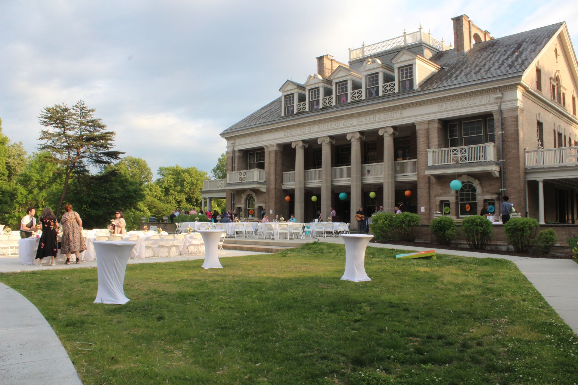 Elegant mansion with columns and lawn lit by evening sun, with guests mingling near white cocktail tables