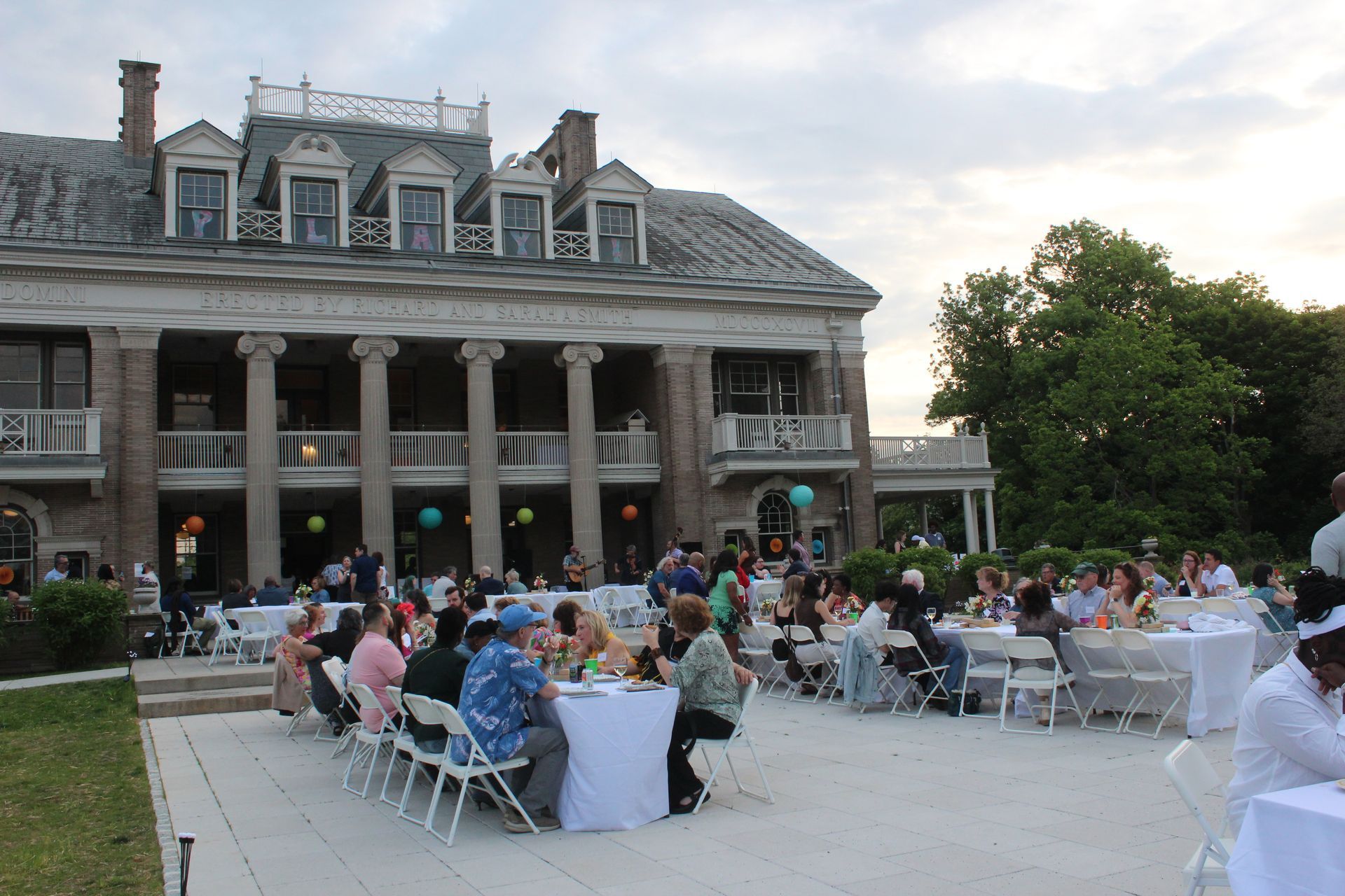 Outdoor wedding reception on a lawn beside a historic mansion, with guests seated at tables and a house with columns in back