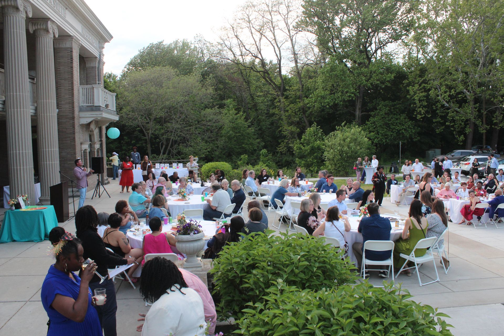 Outdoor dinner party on a patio beside a stone building, with many guests seated at tables near greenery.